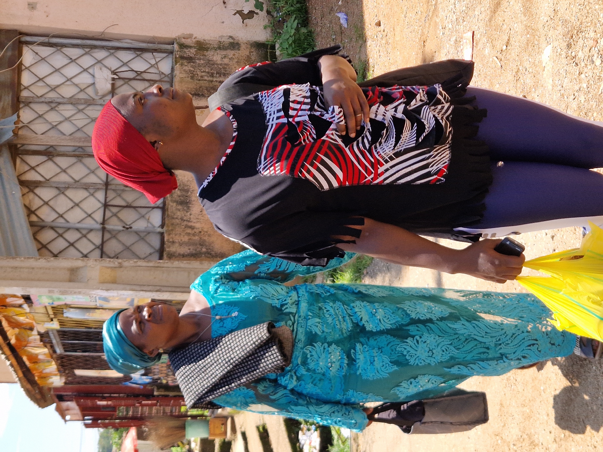 Two women walk down a dusty, unpaved path beside buildings. The woman on the left wears a teal headscarf and a matching teal, long-sleeved lace dress, carrying a dark purse in her left hand and a checkered scarf over her right shoulder. The woman on the right wears a red headscarf and a black t-shirt with a red, white, and black abstract pattern on the front, and dark blue leggings, holding a black smartphone in her right hand and a bright yellow plastic bag in her left. The buildings appear to be shops with barred windows and corrugated metal roofing. The environment suggests a sunny day in an urban area, likely Kabong, Nigeria, characterized by dry, sandy ground and modest structures.