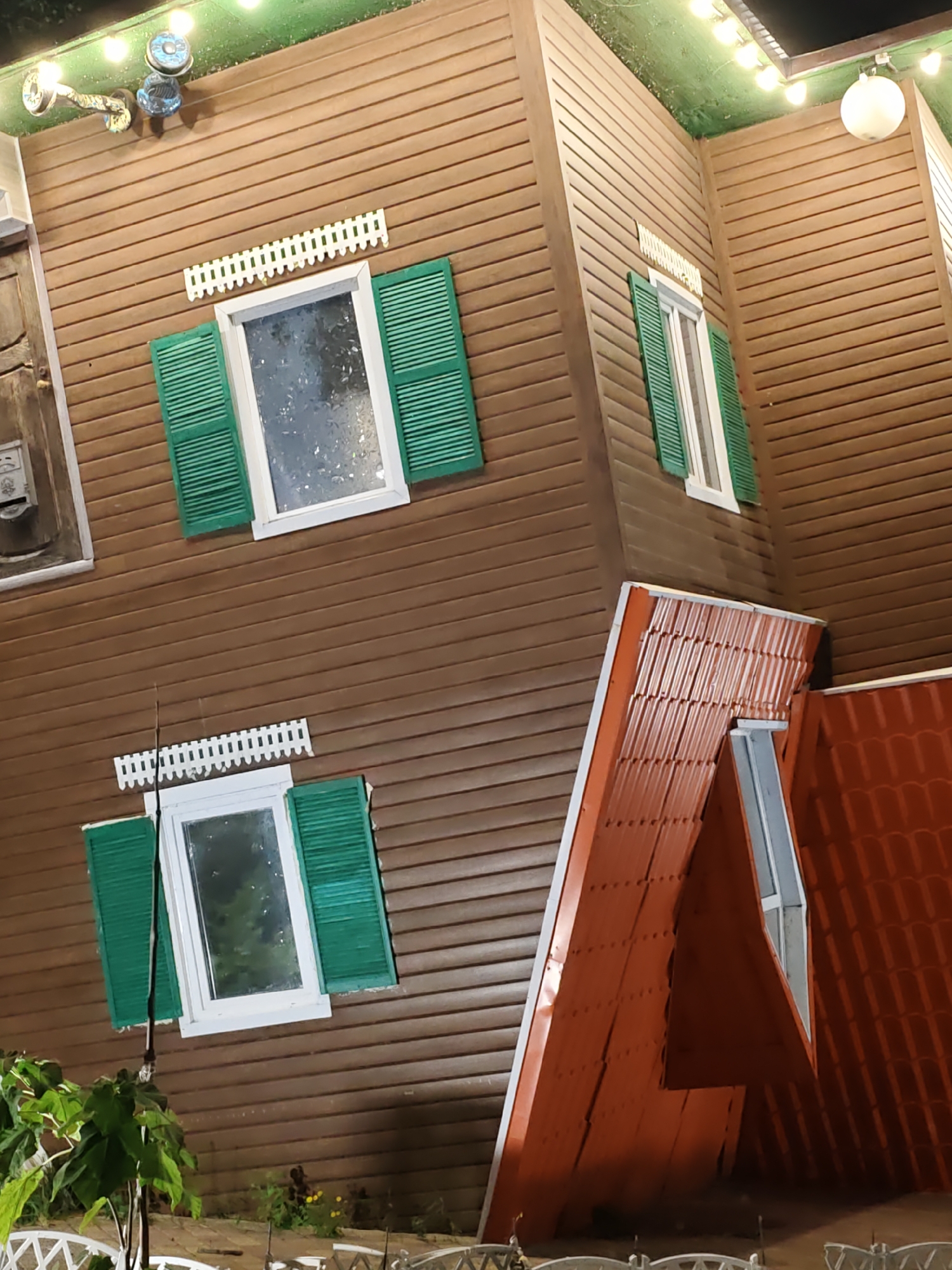 The image shows an upside-down house designed as a tourist attraction, located in Rostov-on-Don, Russia. The house has brown siding and green shutters on its windows. The roof is made of red tiles. Lights are strung along the top of the structure, illuminating the scene. In the foreground, there are some plants and a white picket fence. The overall scene suggests a whimsical and unconventional architectural design.