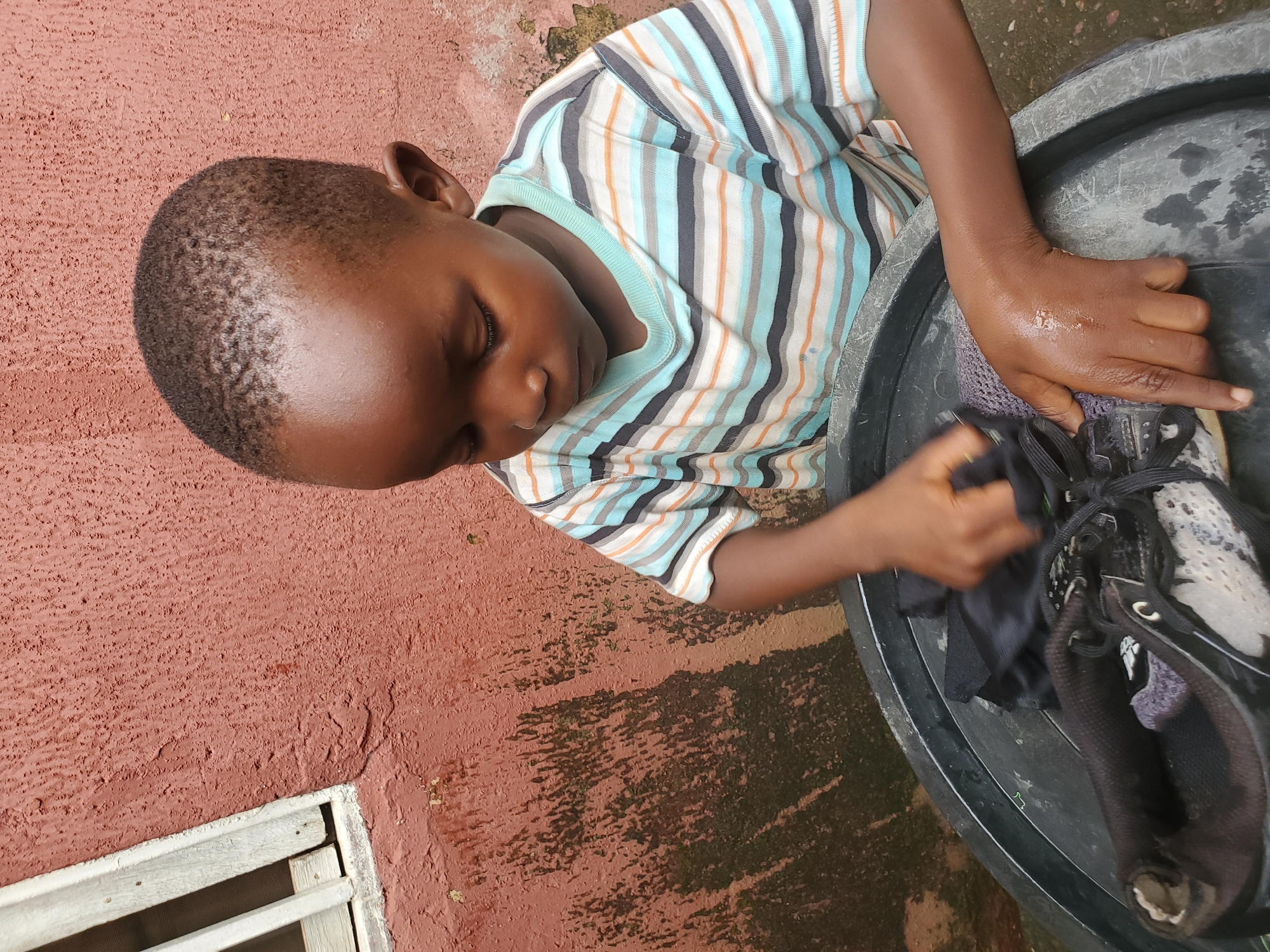A young boy is washing a black and white sneaker in a black plastic basin. He is wearing a horizontally striped t-shirt in shades of blue, orange, gray, and white. The basin is filled with water, and the boy's hands are wet. He is focused on cleaning the shoe, with his head bowed. Behind him is a rough, reddish-pink wall with some patches of green algae. To the left of the boy, a white-trimmed window with vertical white bars is visible against the wall. The scene appears to be outdoors, possibly in an alleyway or yard. The image was taken in Jos, Nigeria.