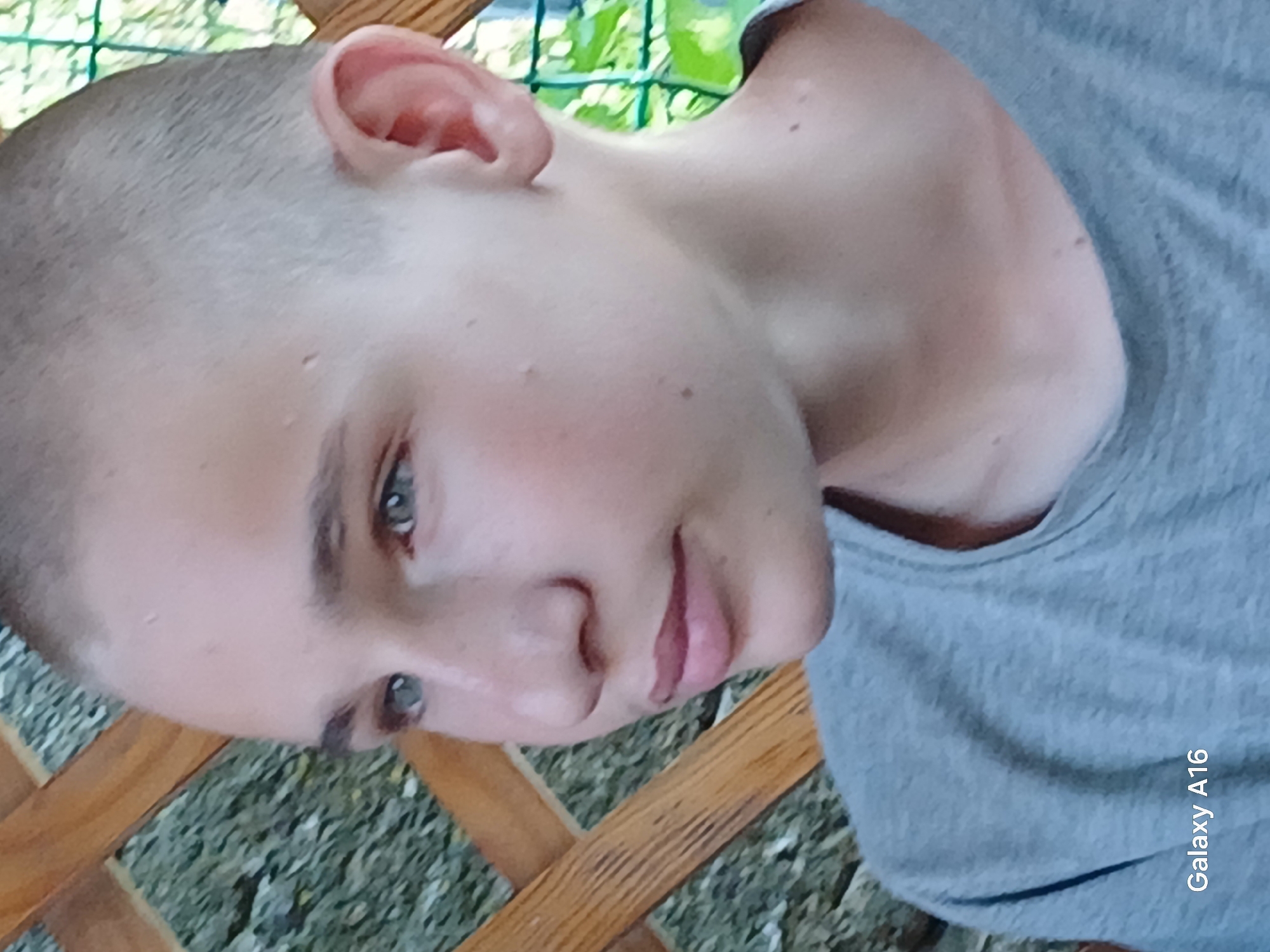 The image is a close-up portrait of a young person with a shaved head, looking to the left. The subject has blue-green eyes and light skin with a few freckles. They are wearing a grey t-shirt. The background appears to be an outdoor setting with a wooden lattice structure and foliage. There is text in the lower left corner that reads "Galaxy A16." The scene is set in Ustyvytsia, Ukraine.