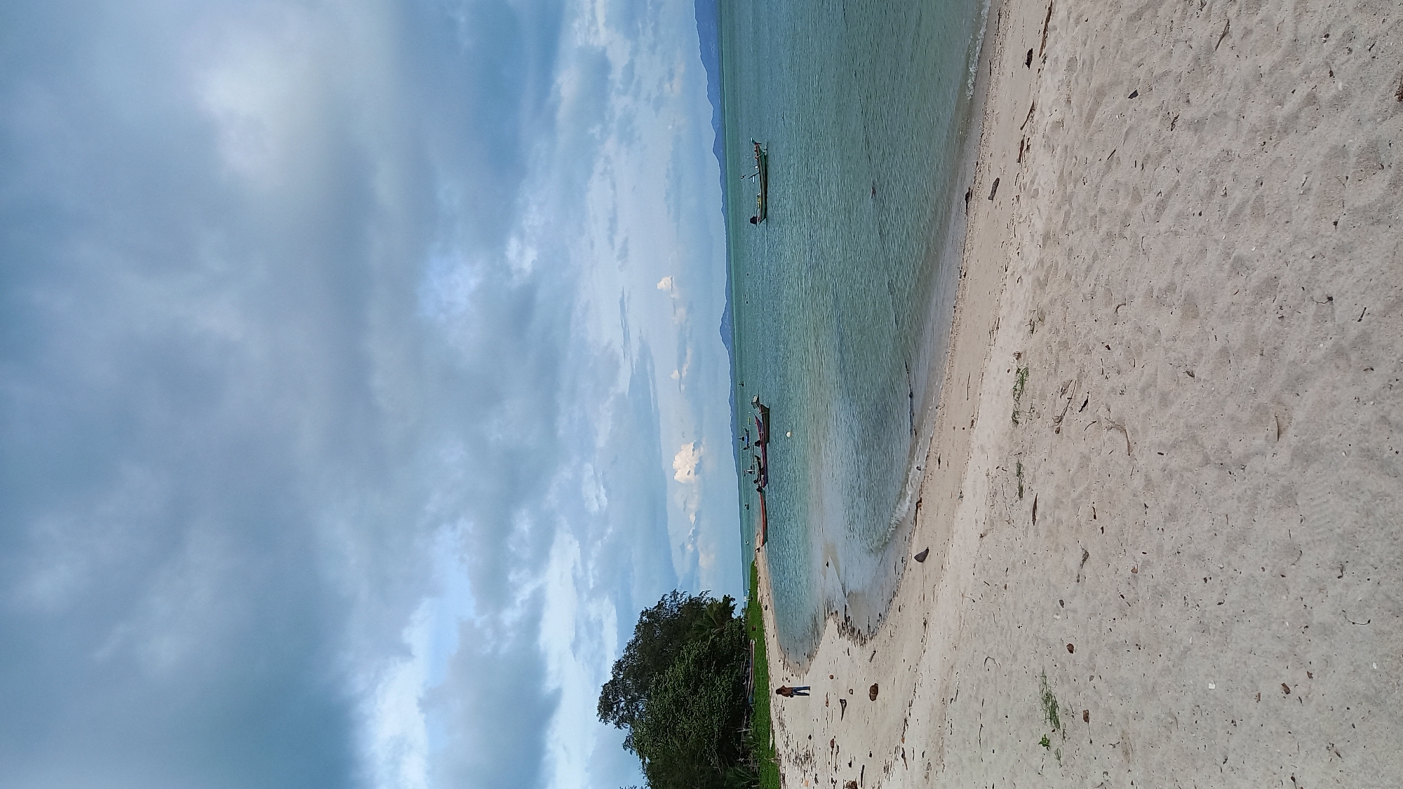 The image depicts a serene beach scene in Ko Pha-ngan District, Thailand. A person is standing on the light-colored sandy beach, facing the turquoise ocean. Several small boats are anchored in the calm water, some clustered together and others more spread out. The background shows a lush green treeline on the left, with distant mountains forming a hazy horizon under a cloudy sky. The overall atmosphere is tranquil and natural.