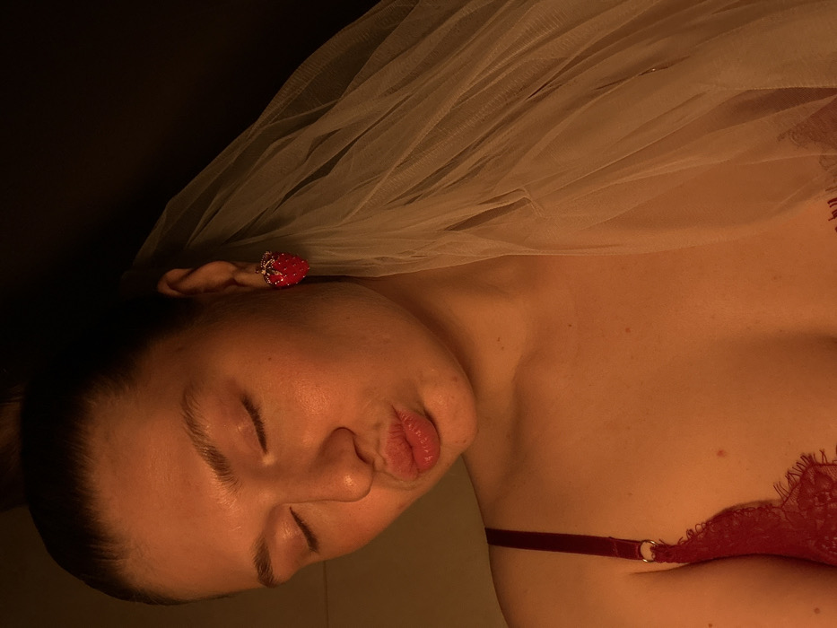 A close-up portrait depicts a single female subject with light skin, her eyes closed, and lips pursed in an expressive gesture. Her head is slightly tilted. Her hair is pulled back, revealing her right ear. She wears a large, red, strawberry-shaped earring with white or silver embellishments on her right earlobe. A sheer, white or light-colored veil or tulle fabric drapes over her right shoulder and partially obscures the right side of the frame. The subject is wearing a red lace bra, with a visible strap and a portion of the lace cup on her left side. The scene is illuminated by warm, directional lighting that highlights her face and upper torso, casting the background into darkness, which lacks discernible features. The specific location within the city of Kazan, Russia, cannot be determined from the visual information present in the image.
