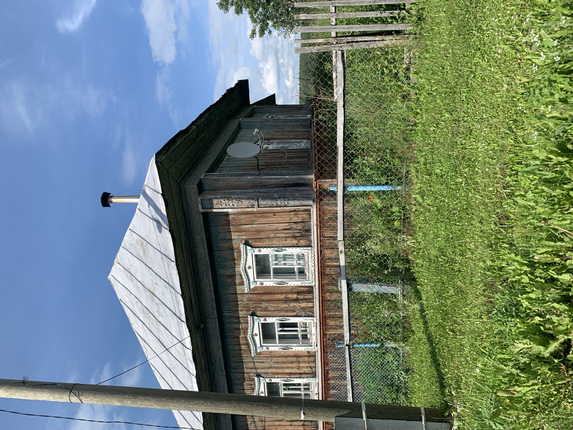 The image shows a weathered wooden house with a tin roof and ornate window trim, set against a blue sky with scattered clouds. A metal satellite dish is attached to the side of the house. In the foreground, a chain-link fence with a wooden section encloses a patch of green grass and overgrown vegetation. A wooden utility pole stands to the left of the house. The scene appears to be in a rural or suburban area.