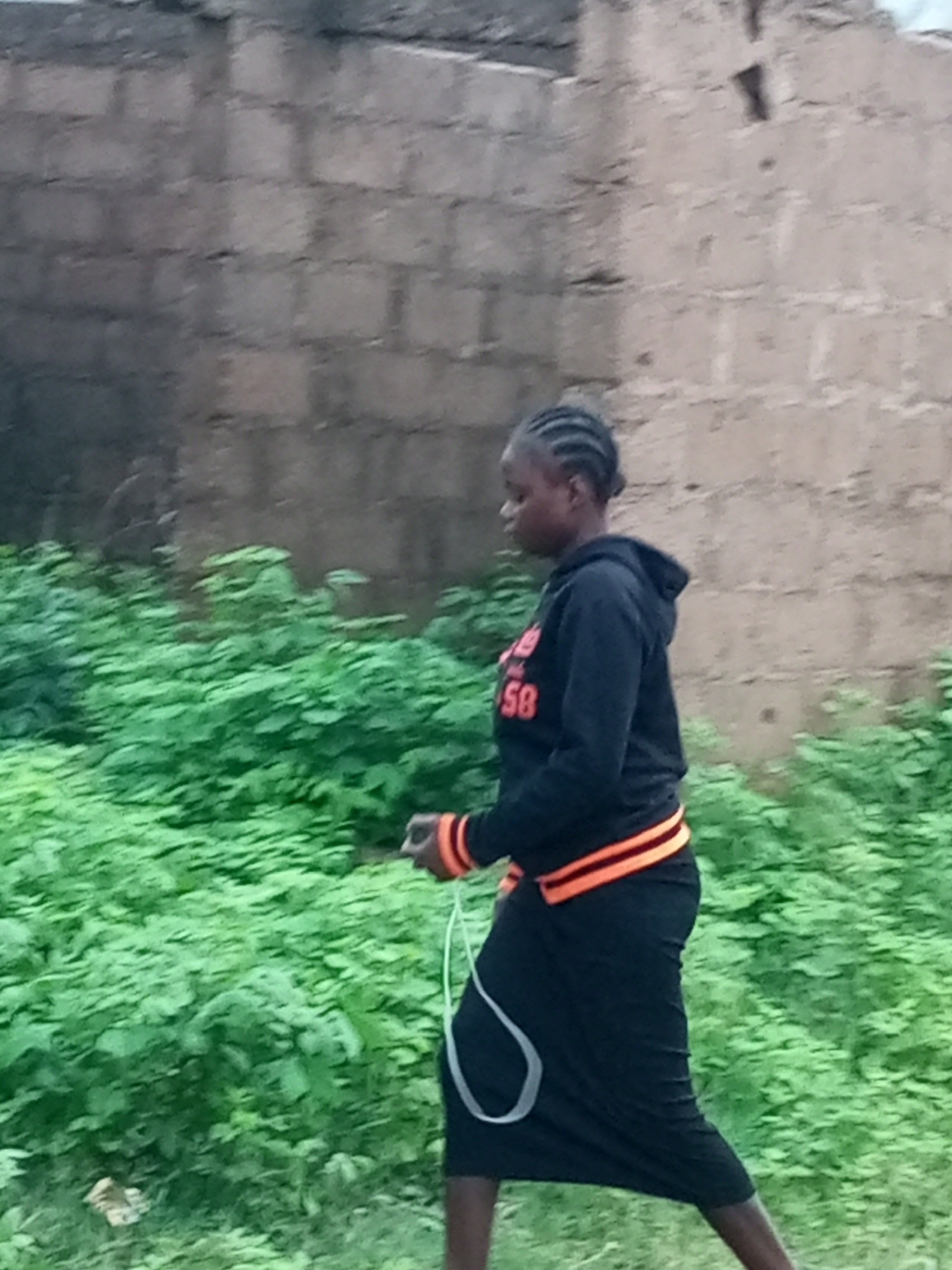 A young woman with braided hair walks in front of a brick wall and lush green foliage. She is wearing a black hooded sweatshirt with orange stripes on the sleeves and waist, and a long black skirt. The woman appears to be walking to the right, and her left hand is visible, holding a white cord. The environment suggests an outdoor urban setting.