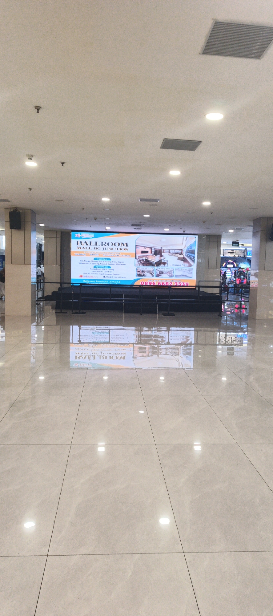The image shows an interior view of a spacious, well-lit venue. A large LED screen prominently displays an advertisement for "BALLROOM MALL BG JUNCTION", advertising its suitability for weddings, seminars, and parties, along with a phone number and room details. The screen is mounted on a stage with black curtains. To the right of the screen, there are some arcade-style game machines visible, and a glass partition separates the ballroom area from another space. The floor is polished and reflective, with visible lines from large tiles. The ceiling features recessed lighting and air vents. The overall environment suggests a modern event or shopping mall complex. The reflection on the floor shows a reversed image of the LED screen.