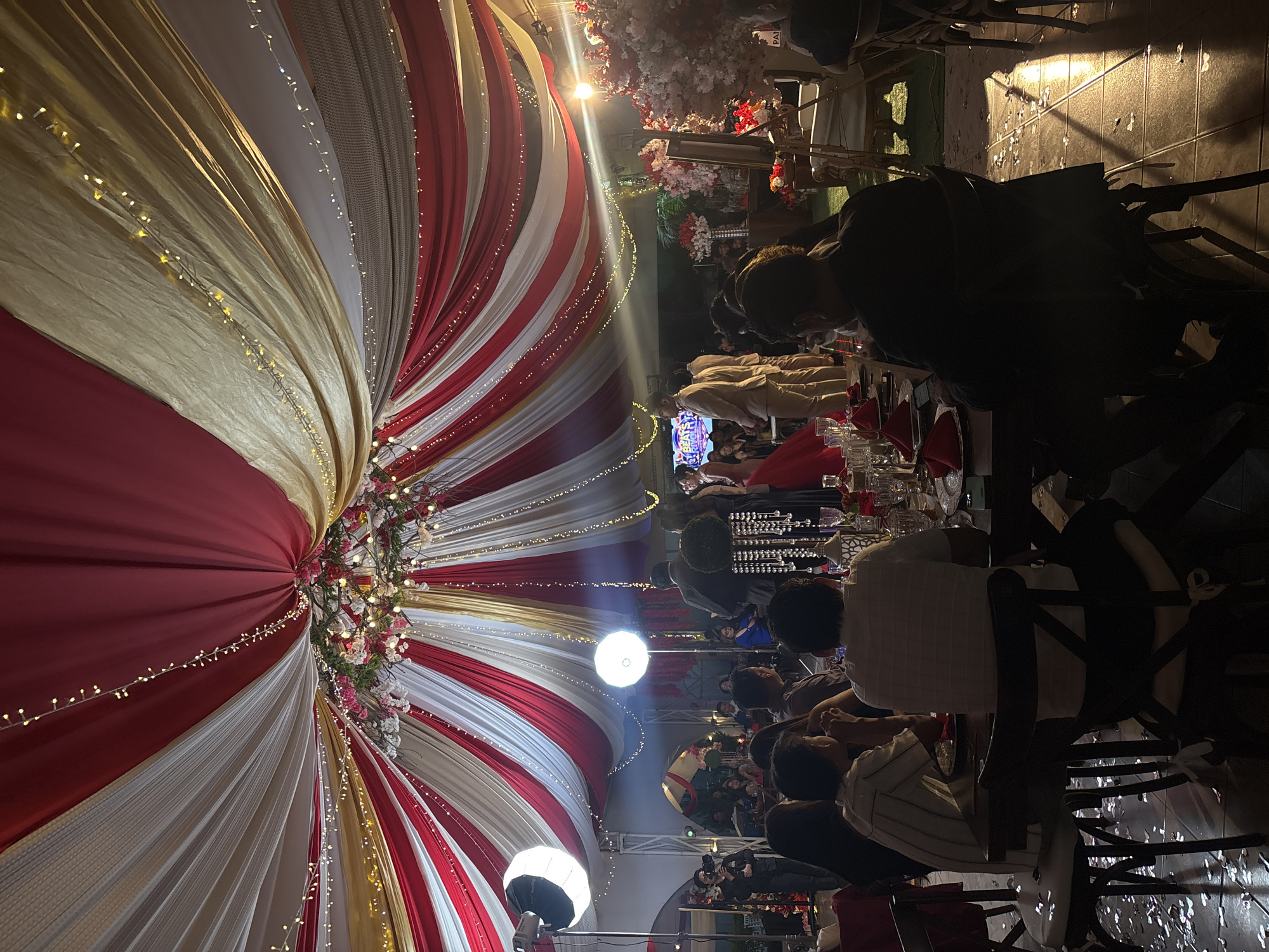 The image depicts a festive event, possibly a wedding reception, under a large tent with red, white, and gold draping. The tent is decorated with strings of lights and floral arrangements. Tables are set with dinnerware and red napkins, where seated guests are seen engaged in conversation or eating. The lighting is a mix of natural and artificial sources, with prominent spotlights and ambient light casting shadows across the scene. The location is unspecified, but the city of Chilpancingo, Mexico is mentioned as a possible location. People are seated at tables and some are standing, engaged in conversations and enjoying the event.
