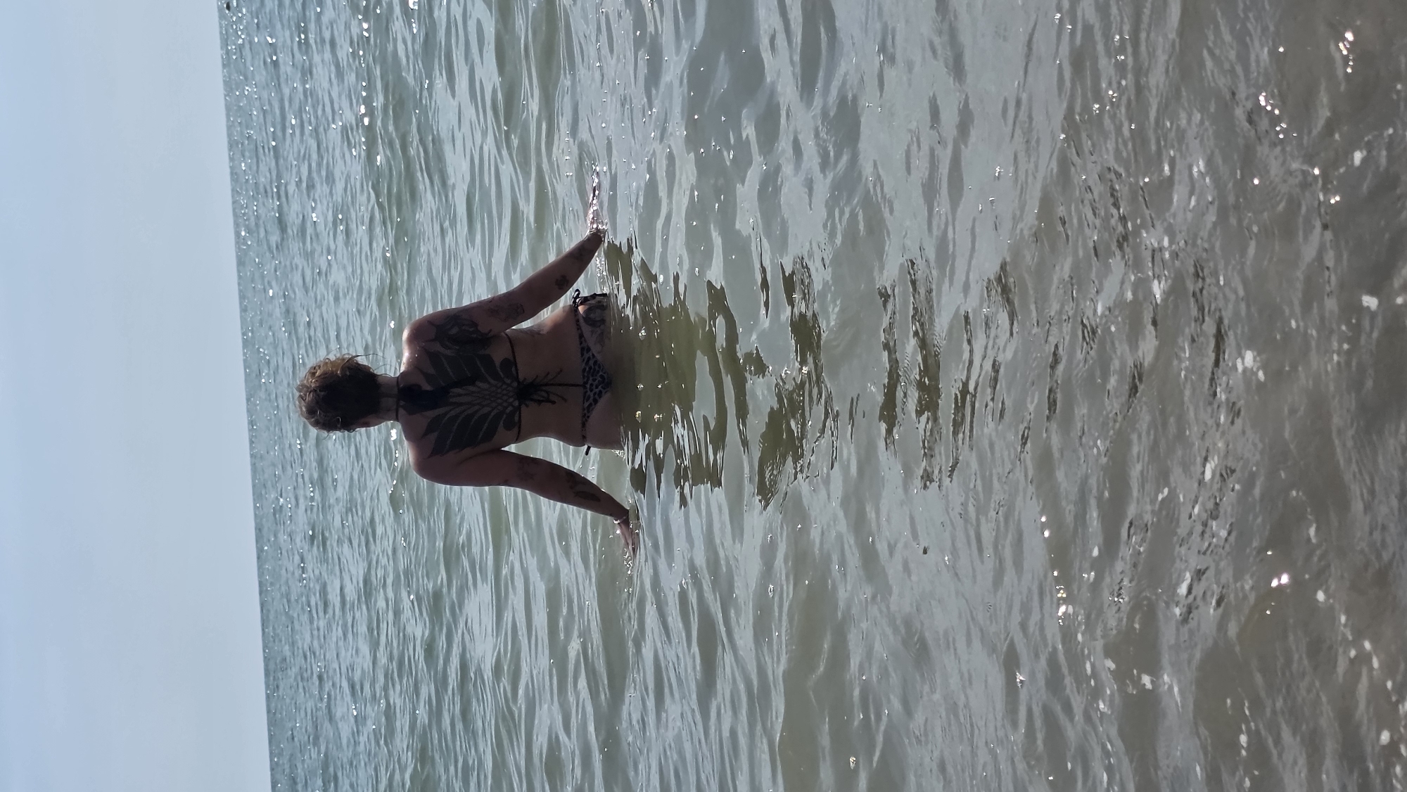 A person wearing a black bikini stands in shallow ocean water, facing away from the camera. They have a large, black, wing-like tattoo on their back and smaller tattoos on their arms. The water is calm with small ripples, and the sky is clear and blue. The scene appears to be a beach.