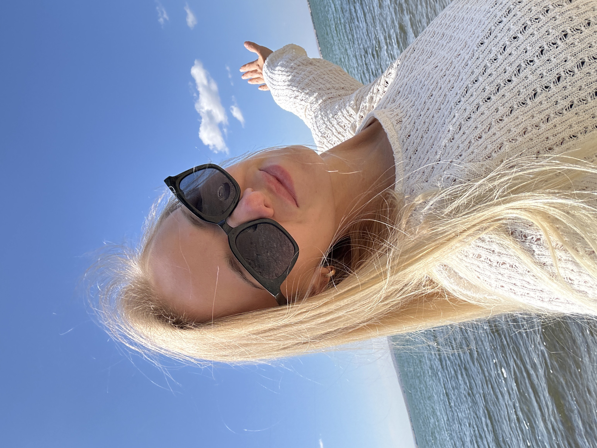 A woman with blonde hair and sunglasses is taking a selfie outdoors. She is wearing a white knitted sweater and her right arm is raised, with her palm facing outward. She is standing on the shore of a body of water, which appears to be a lake or a bay, with gentle waves. The sky is blue with a few scattered clouds. The background shows a distant shoreline. The overall scene suggests a sunny day in a natural setting, possibly near the city of Gornyi Erik, Russia, given the context.