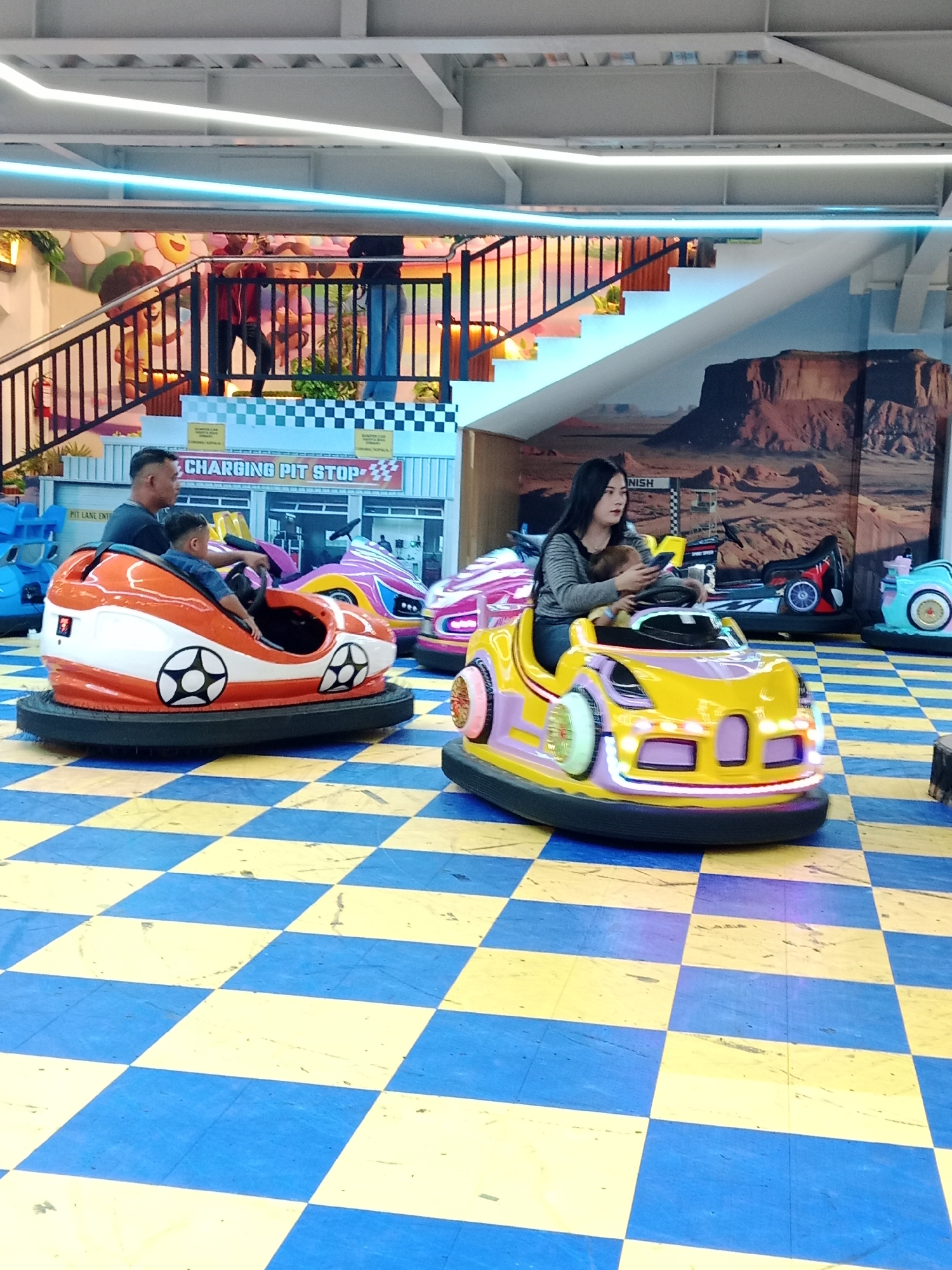 This is a medium shot, eye-level photograph of a bumper car attraction with people inside. The attraction is located at a shopping mall in Kecamatan Pati, Indonesia. The floor is covered in a yellow and blue checkered pattern. In the foreground, a man is sitting in an orange and white bumper car with a child in front of him. In the middle ground, a woman is sitting in a yellow and purple bumper car with a child in front of her. In the background, there are more bumper cars and a staircase. The overall lighting is bright and even.