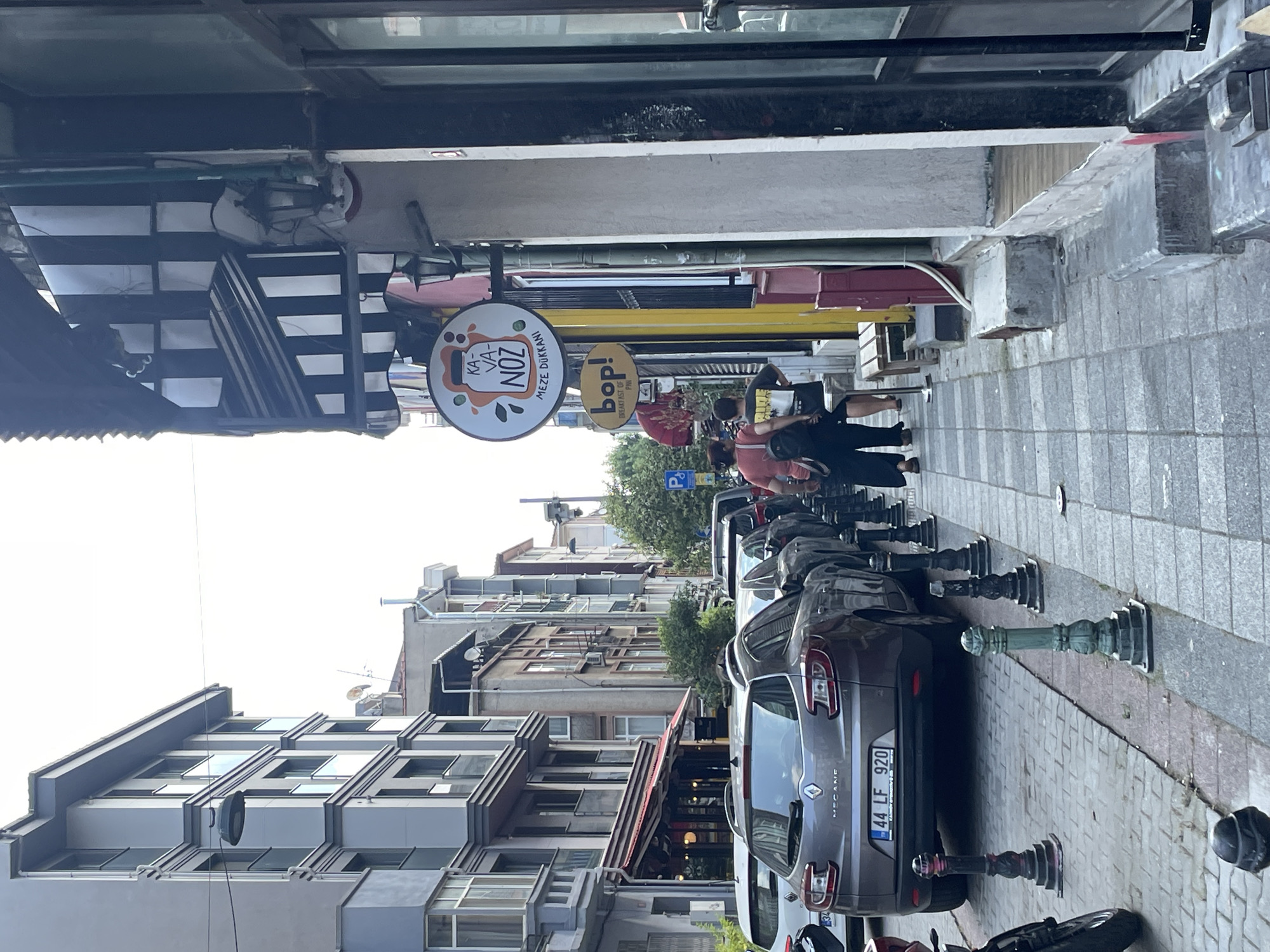 In Kadıköy, Turkey, a street scene unfolds with buildings lining both sides. On the left, a modern gray building with multiple floors and balconies dominates the view. On the right, a narrower sidewalk runs alongside a row of buildings with awnings and storefronts. Several cars are parked along the street, including a gray Renault Megane in the foreground with license plate "44 LF 920". Two people, a woman in an orange shirt and a man in a black t-shirt, walk away from the camera on the sidewalk. Above them, signs for "KAVA NOZ MEZE DÜKKANI" and "bop!" are visible, indicating commercial establishments. The street is paved with cobblestones and has decorative bollards lining the sidewalk. The sky is overcast, and the overall atmosphere suggests a typical urban environment in Turkey.