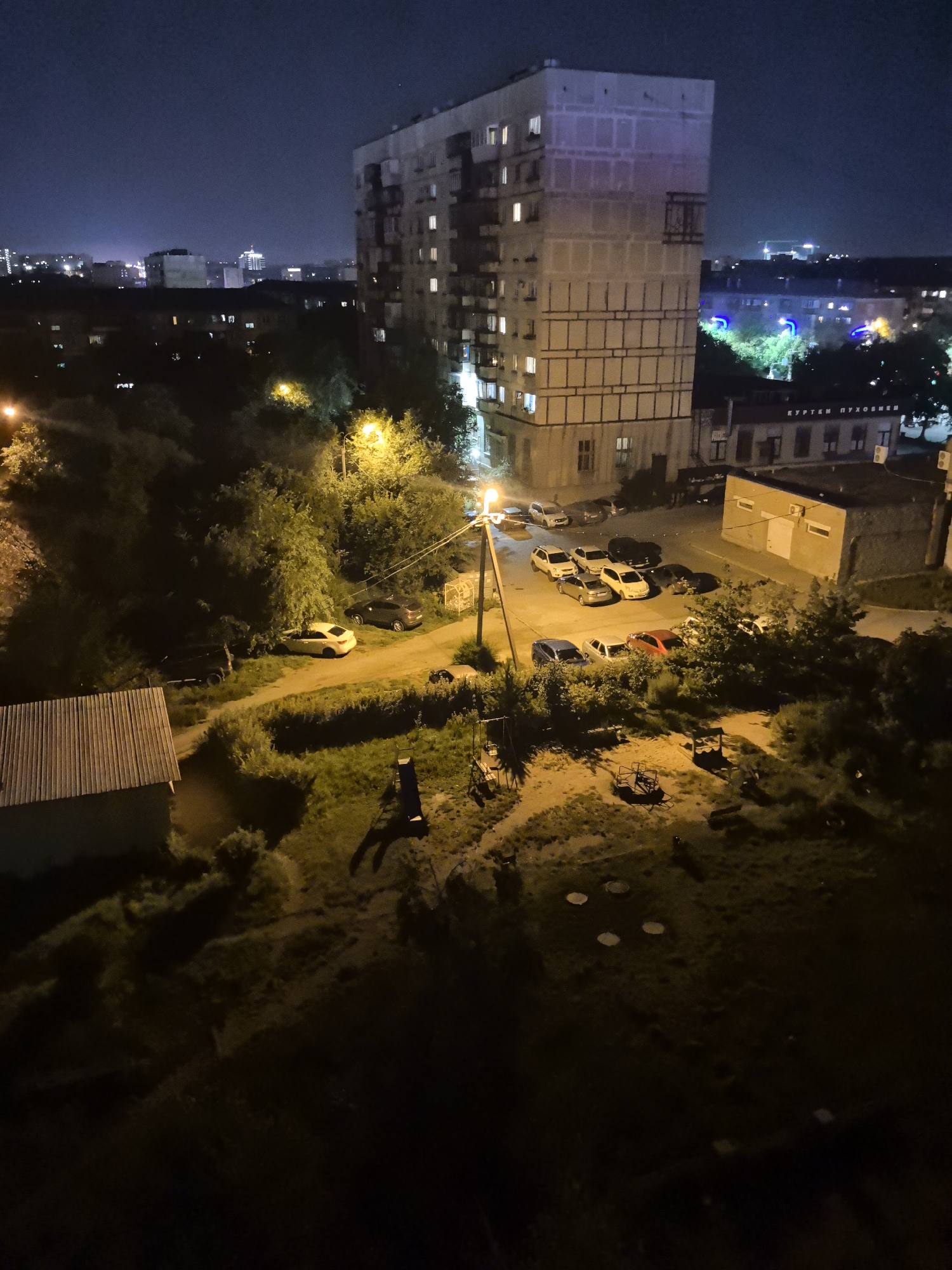 The image depicts an outdoor nighttime scene in an urban environment, specifically identified as being in the city of Magnitogorsk, Russia. The foreground is dominated by dark, unlit ground with patches of grass and some indistinct playground equipment. A small, light-colored building with a corrugated metal roof is visible on the left side.
Further into the scene, a paved area or road is illuminated by streetlights, revealing a number of parked cars. These include several sedans and SUVs, in various colors such as white, silver, black, and red. Trees with dense foliage are interspersed throughout the area, some lit by the streetlights.
In the mid-ground, a multi-story apartment building stands prominently. It is rectangular in shape with visible windows, some of which are illuminated from within. The facade of the building appears to be a light concrete or stucco material. Adjacent to the apartment building is a smaller, single-story structure with a flat roof and minimal visible features.
In the background, other buildings and structures are visible, suggesting a densely populated urban area. Some of these buildings have illuminated windows or lights. The sky above is dark, indicative of nighttime, with a faint glow from distant city lights. The overall atmosphere is one of a typical residential area at night, with artificial lighting casting shadows and highlighting certain features.