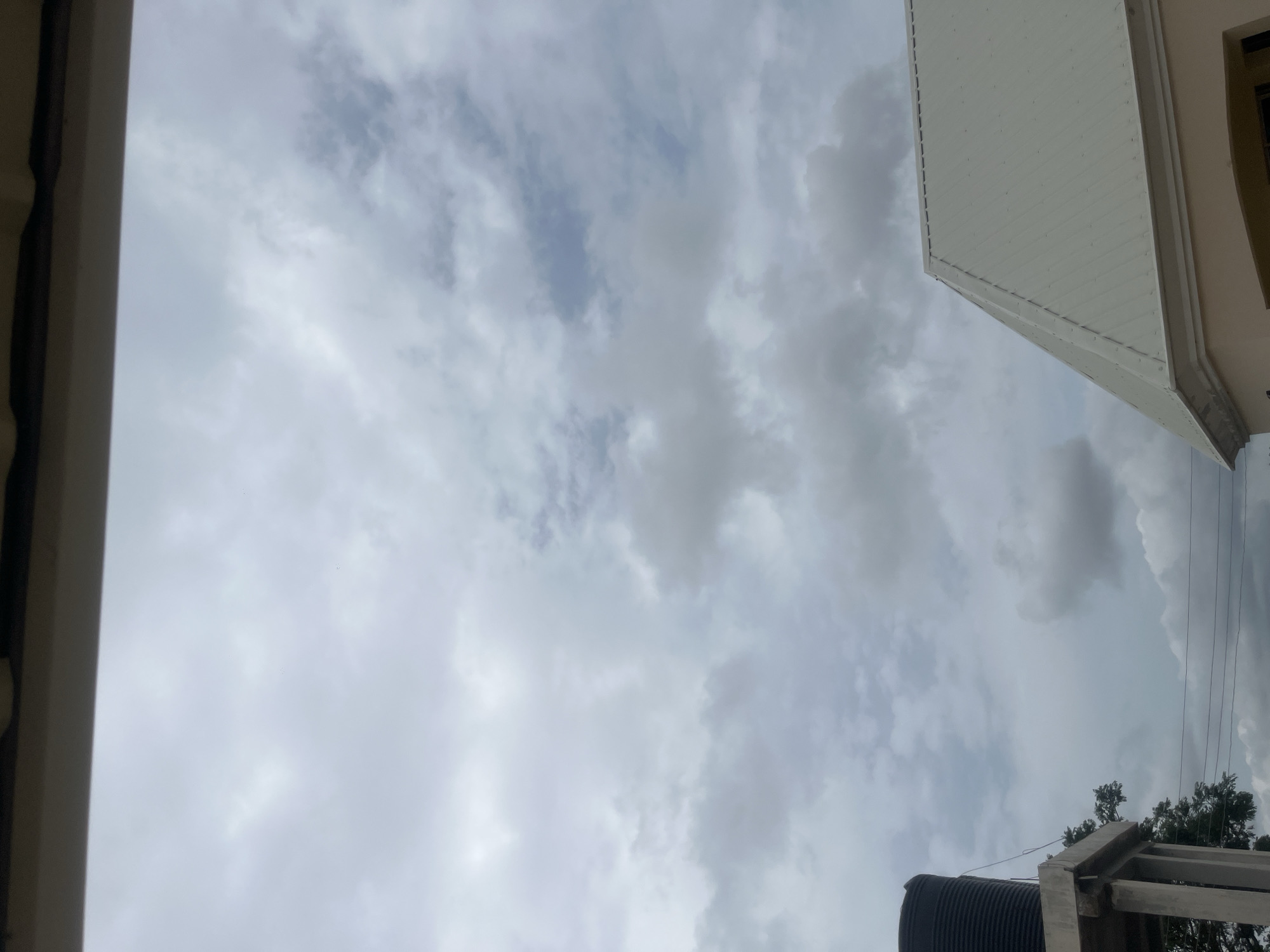 The image shows a cloudy sky above a rooftop and a water tank in Jauro Yinu, Nigeria. A portion of a beige building with a white metal roof is visible in the lower right corner. To the left, a black water tank is mounted on a concrete platform supported by pillars, with some tree foliage visible behind it. Electrical wires crisscross the scene. The sky is overcast with grey and white clouds.
