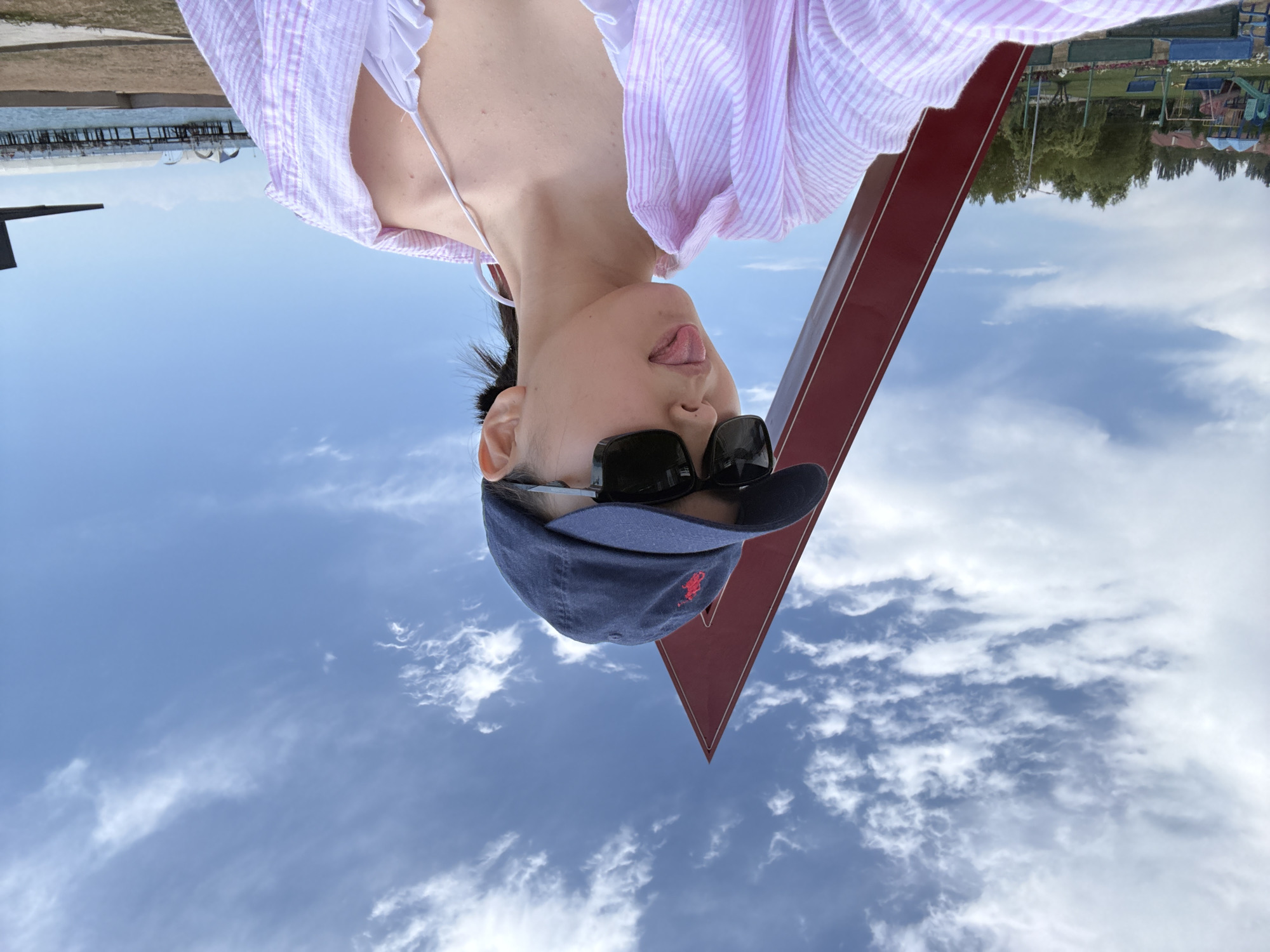 An outdoor selfie captures a young woman wearing a blue baseball cap and sunglasses, sticking her tongue out playfully. She is dressed in a pink and white striped unbuttoned shirt over a white bikini top. Behind her, a large, red triangular sculpture stands against a bright blue sky with scattered clouds. In the background, a playground with slides and swings is visible, along with trees and buildings. Further beyond, a pier extends over a body of water, and a distant structure with arches is seen. The scene appears to be in the city of Sary-Oy, Kyrgyzstan.