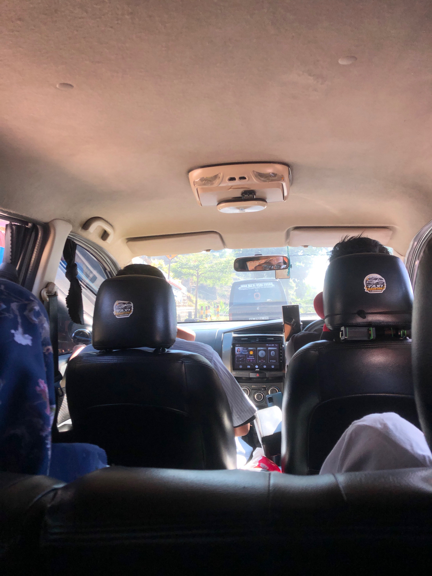 The image is an interior view of a taxi in Dadi, Indonesia, looking forward from the back seat. The taxi is a modern sedan, likely a Nissan Grand Livina, with black leather seats. Two passengers are visible in the back, one with a blue and white patterned shirt on the left, and another with dark hair and a white cloth on the right, partially obscured. The driver's headrest has a sticker that reads "TAXI MANDARIKARYA." The dashboard features a large touchscreen infotainment system, and a smartphone is mounted on a stand. Outside the windows, a street scene is visible with trees and another vehicle. The lighting suggests it is daytime.