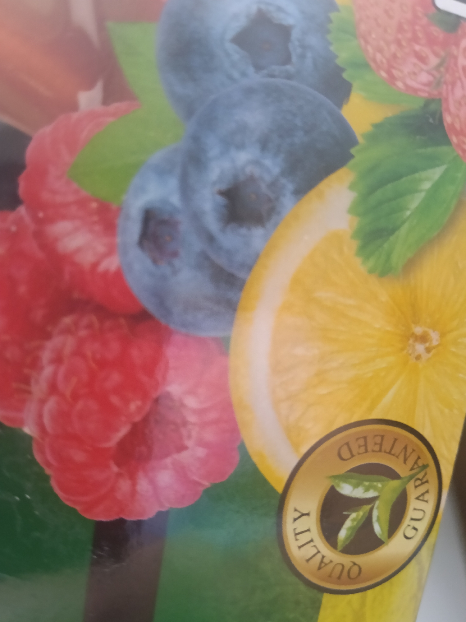 The image shows a close-up of a packaging design featuring various fruits. Prominently displayed are blueberries, raspberries, and a slice of lemon. There is also a round golden seal with the words "QUALITY GUARANTEED" and an image of tea leaves. The background is a gradient of green and yellow. There is no indication of the city of Altinkul, Uzbekistan, in this image.
