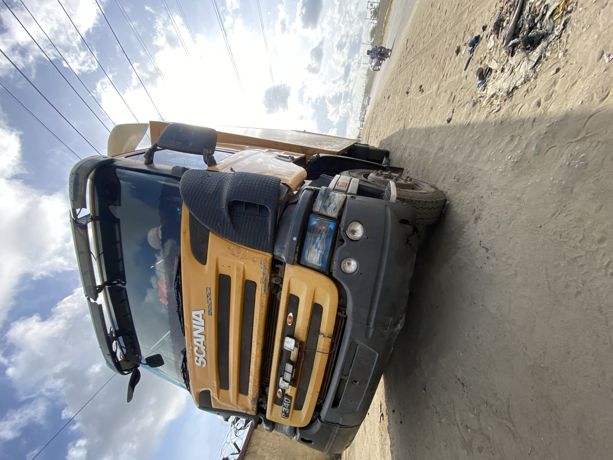 The image shows a yellow and black Scania P 340 truck parked on a dirt road. The truck is facing the camera, with the driver's side slightly visible. The word "SCANIA" is prominently displayed on the yellow front panel. Overhead power lines are visible against a cloudy sky. In the background, there is a motorcycle with two people on it traveling away from the camera on the dirt road, along with trash and debris strewn to the right. The setting is identifiable as Lekki, Nigeria.
