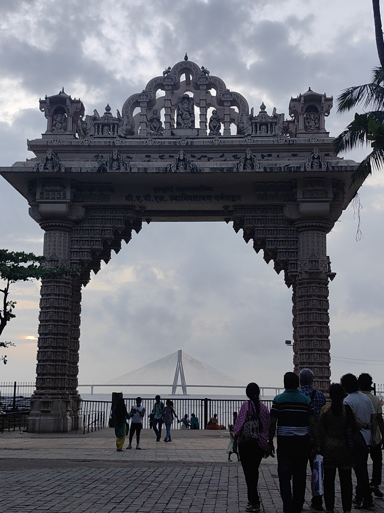 This image captures a view through a large, ornate stone archway, likely a gateway to a temple or significant cultural site in Mumbai, India. The arch is intricately carved with statues and decorative motifs, typical of Indian temple architecture. In the background, visible through the arch, is a prominent suspension bridge, which is characteristic of the Bandra-Worli Sea Link in Mumbai. The sky is overcast, with clouds filling the frame. Several people are walking on a paved walkway in front of the archway, appearing to be tourists or locals enjoying the area. The overall scene suggests a blend of cultural heritage and modern infrastructure in a coastal urban environment.