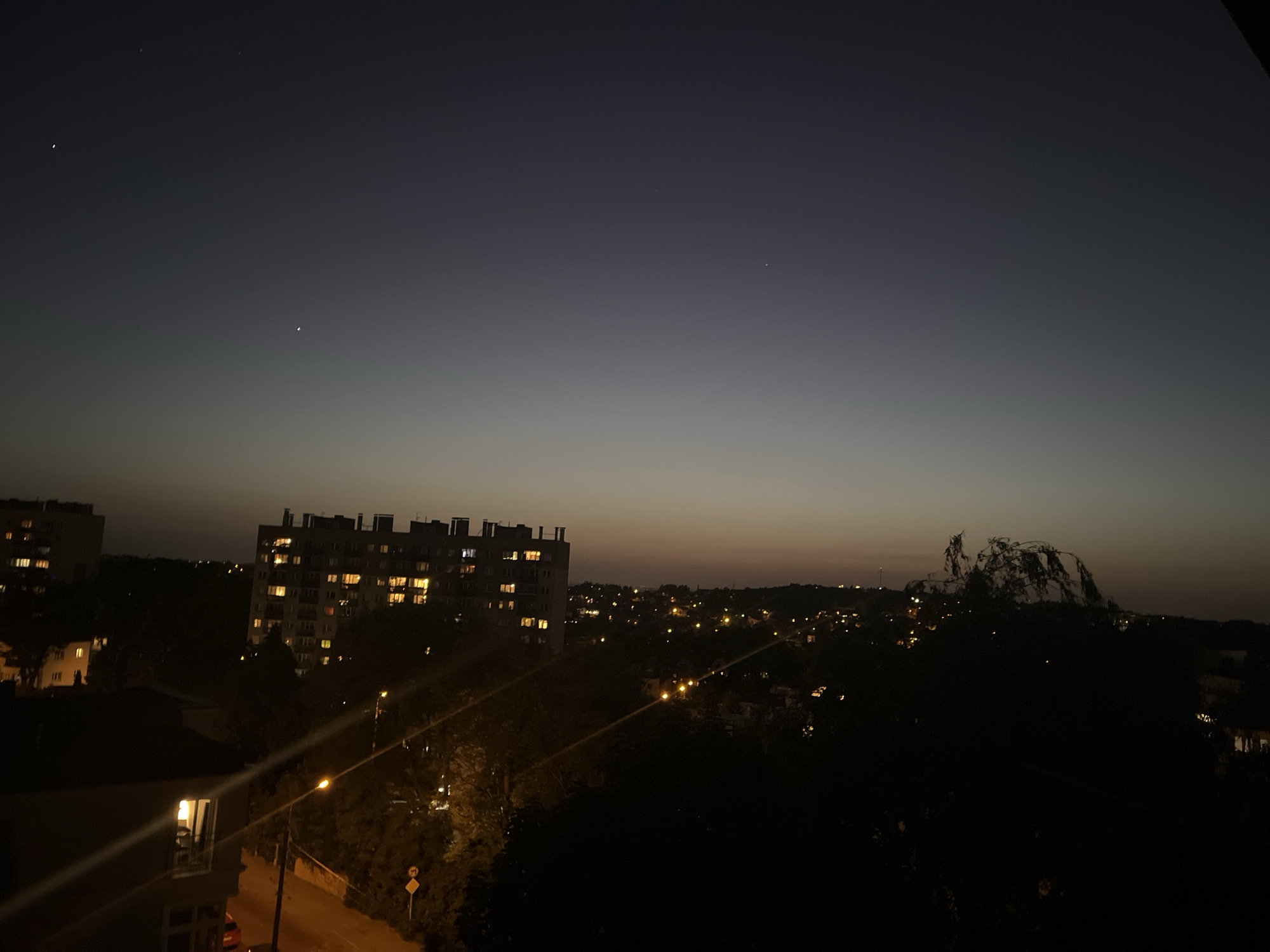 The image depicts a cityscape at night or twilight in Unknown, Unknown. Several multi-story buildings with illuminated windows are visible in the middle ground. Streetlights line the roads, casting a warm glow on the surroundings. Dark trees fill the foreground, obscuring some of the view. The sky is a gradient of dark blue to lighter hues near the horizon, with a few stars visible.
