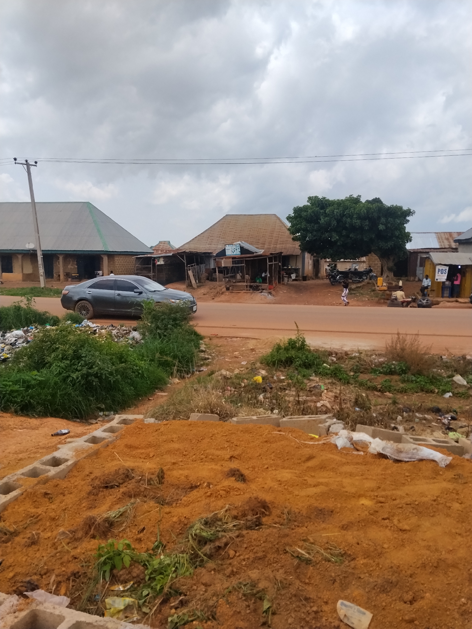 The image depicts a street scene in Durumi, Nigeria, under a cloudy sky. A dark gray sedan is driving on a reddish-brown paved road. To the left of the road, there is a large pile of green vegetation and garbage, with some concrete blocks in the foreground. Across the street, there are several buildings with corrugated metal roofs. One building has a sign that appears to read "POS" and has people gathered in front of it. Another building, further to the left, has a similar roof and appears to be a shop or residence. A large green tree stands behind some of the buildings. The ground is uneven and appears to be dirt and gravel. A young child is walking on the road.