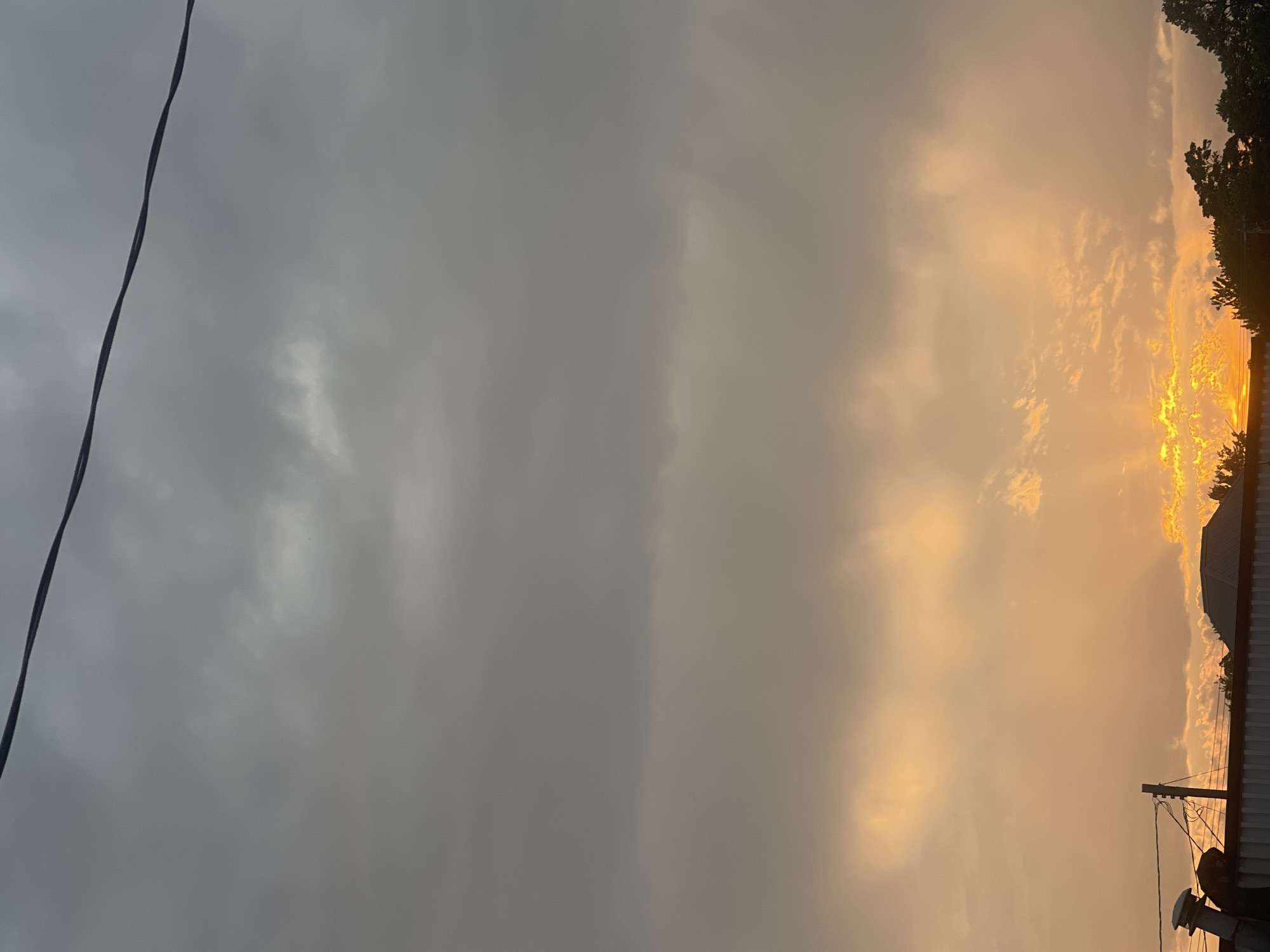 The image captures a cloudy sky during sunset, with the sun's rays breaking through the clouds in a vibrant display of orange and yellow light. The lower portion of the image shows the silhouette of a suburban landscape, featuring rooftops, trees, and electrical wires, possibly indicative of a residential area within Bluewater, Canada. The atmosphere appears overcast, suggesting potential for rain or impending weather changes. No individuals or direct human interaction are visible in the scene.
