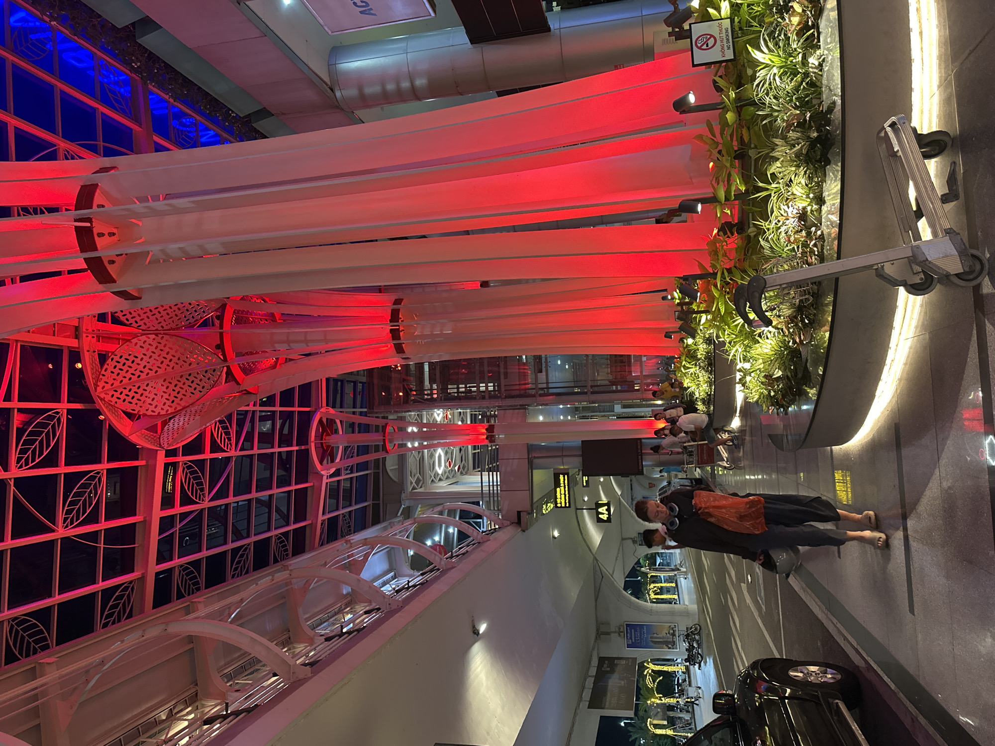 The image shows the exterior of Da Nang International Airport in Da Nang, Vietnam, at night. The architecture features modern design elements with extensive use of glass and red lighting. A woman is walking on the sidewalk towards the camera, carrying a shopping bag and a rolling suitcase. She is wearing dark clothing and a pair of headphones around her neck. In the background, several cars and motorcycles are parked, and other people are visible walking and waiting. The airport's interior lighting, with its red and blue hues, reflects on the wet pavement, suggesting recent rain. Lush green plants are strategically placed around the building's pillars, adding a natural element to the urban landscape. A luggage cart is parked in front of the plants. The overall impression is one of a busy and well-lit transportation hub at night.