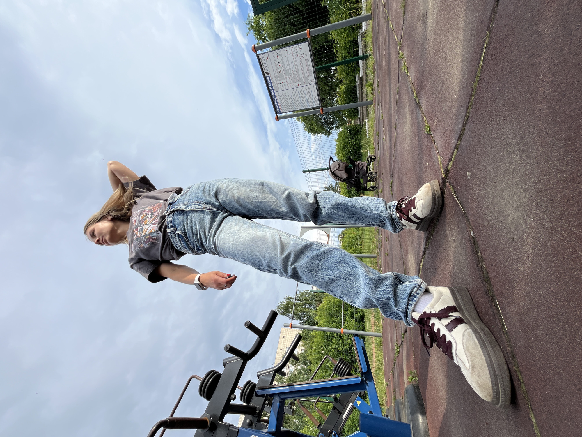 A low-angle outdoor shot shows a young woman standing on a red rubberized surface in a park, possibly in Chaikovsky, Russia, given the presence of a sign with Russian text. She is wearing a grey t-shirt with a graphic print, light blue distressed jeans, white socks, and beige and maroon athletic shoes. Her right arm is raised, with her hand behind her head, and she is looking upwards and to her right. To her left is a blue and black weight training machine. In the background, a fence encloses an area with greenery, and a baby stroller is visible. A large informational sign is mounted on a pole behind the stroller. The sky is overcast with white clouds.