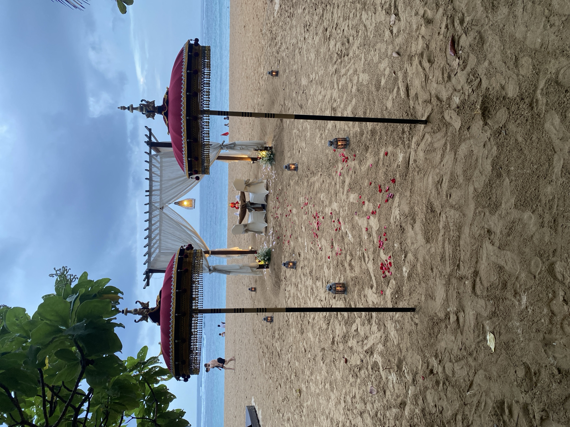 A romantic dinner setup is arranged on a sandy beach in Kuta Selatan, Indonesia. The scene features a small, elevated wooden structure with white drapes, under which a table for two is set with red napkins and what appears to be a candle centerpiece. Surrounding the table are two chairs, and a pathway of scattered rose petals leads towards it. Two ornate red and gold parasols stand prominently in the foreground, flanking the path. Several small lanterns are placed along the sand, adding to the ambiance. In the background, the ocean meets a cloudy sky, and a few people are visible walking and sitting on the beach. Lush green foliage frames the left side of the image. The lighting suggests it is either early morning or late evening.
