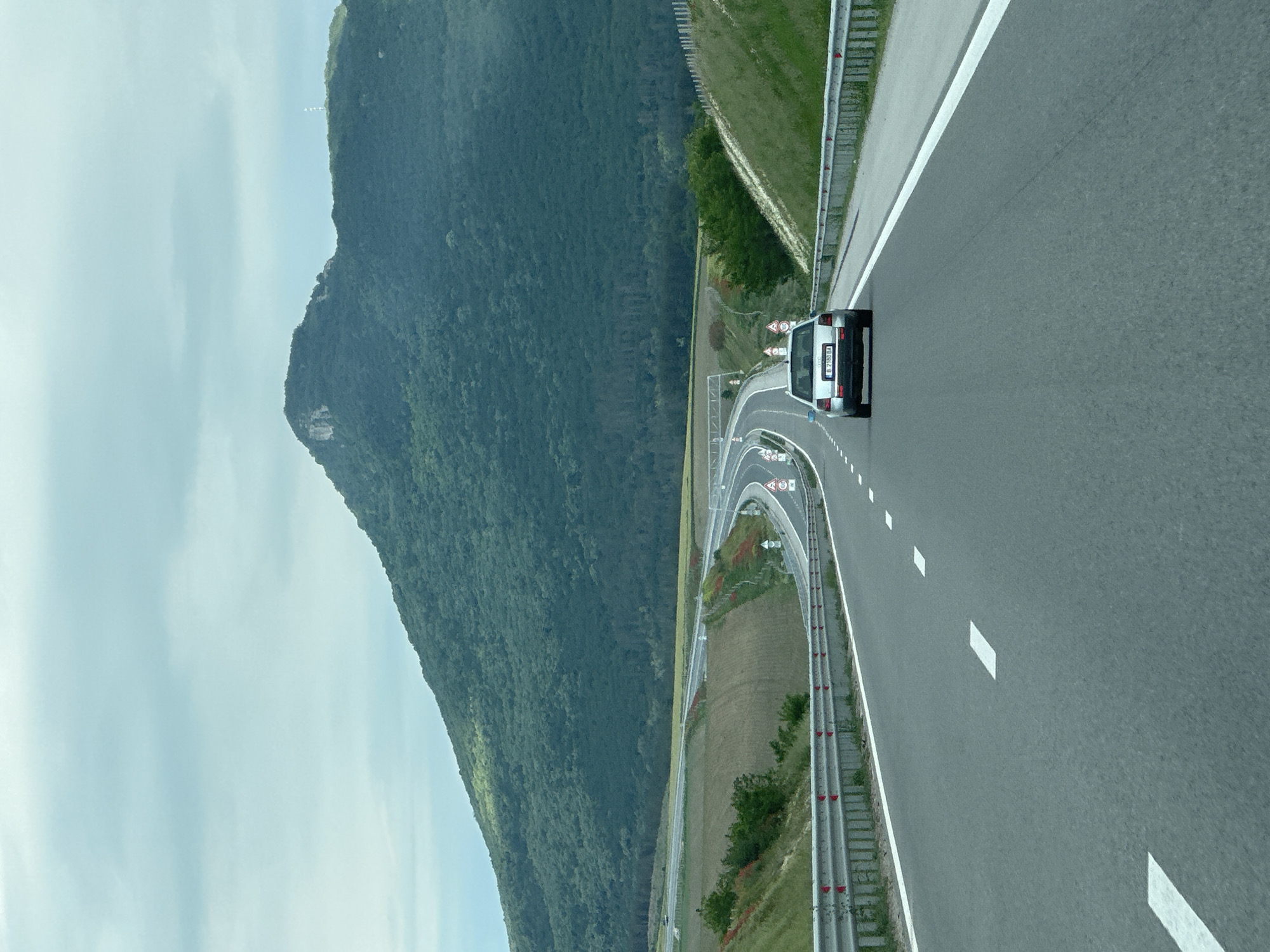 The image shows a silver vehicle driving on a highway near Buhovtsi, Bulgaria. A mountain with dense vegetation dominates the left side of the frame. The highway curves with several warning signs visible. The sky is overcast with a faint blue hue. The road has lane markings and guardrails on both sides.
