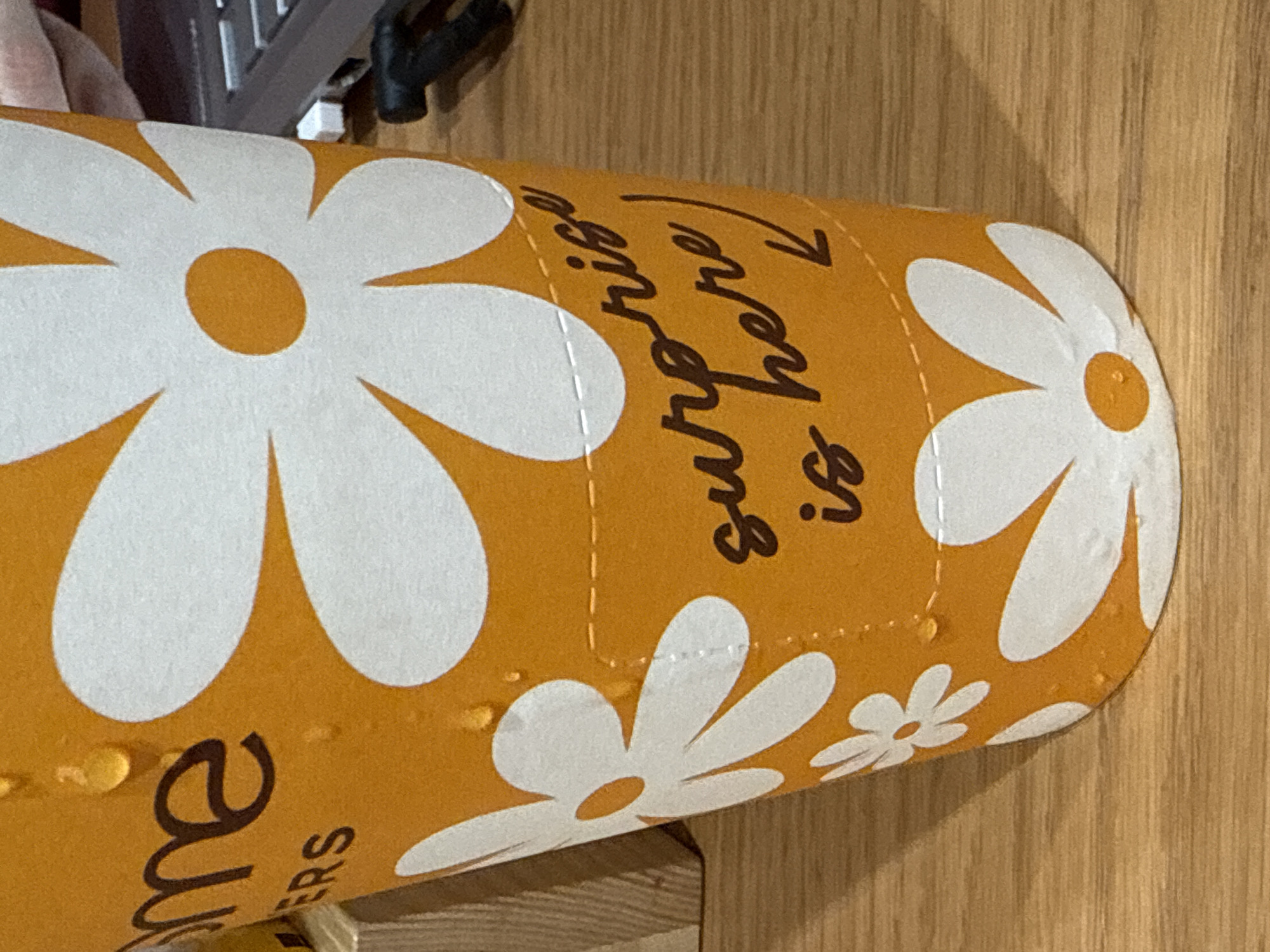 The image displays a close-up of an orange cylindrical object, likely a drink container, resting on a light-colored wooden surface. The object is decorated with a pattern of white daisy-like flowers, each featuring an orange center. Numerous small, clear water droplets are visible on its surface, indicating condensation.
Dark brown cursive text is printed on the object. On the right side, the words "surprise is here" are clearly visible, accompanied by a downward-pointing arrow. On the far left, partially obscured, the text "me" is visible, with "ERS" printed below it, suggesting part of a larger word.
The background is soft and out of focus, showing indistinct dark shapes that may be other objects or furniture. The wooden surface displays a subtle grain pattern. The scene is illuminated by ambient light, suggesting an indoor setting. No individuals or active events are depicted.