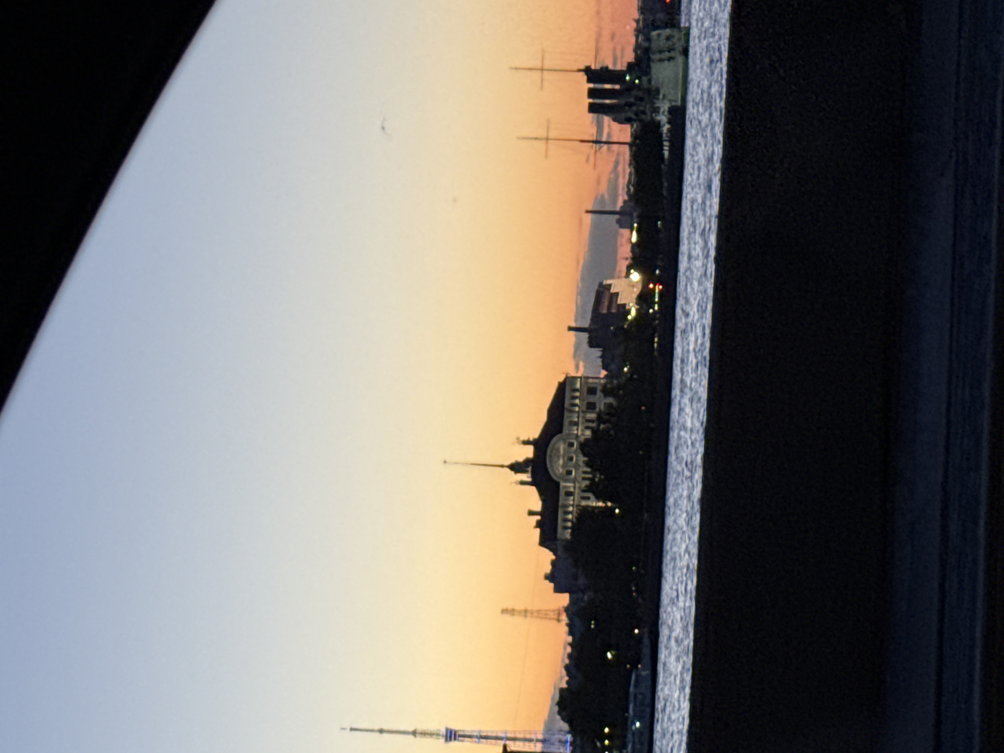 The image depicts a waterfront view of Saint Petersburg, Russia, during sunset. In the background, silhouetted against the orange and blue sky, are several prominent structures. To the left, a tall, slender radio tower with lights is visible. To the right of that, a complex of buildings with a distinctive spire crowns the skyline. Further to the right, a warship with prominent masts and gun turrets is docked or anchored. The foreground shows a dark, solid structure, possibly a wall or embankment, overlooking a body of water that reflects the fading light of the sky. Scattered lights from the city can be seen along the waterfront.