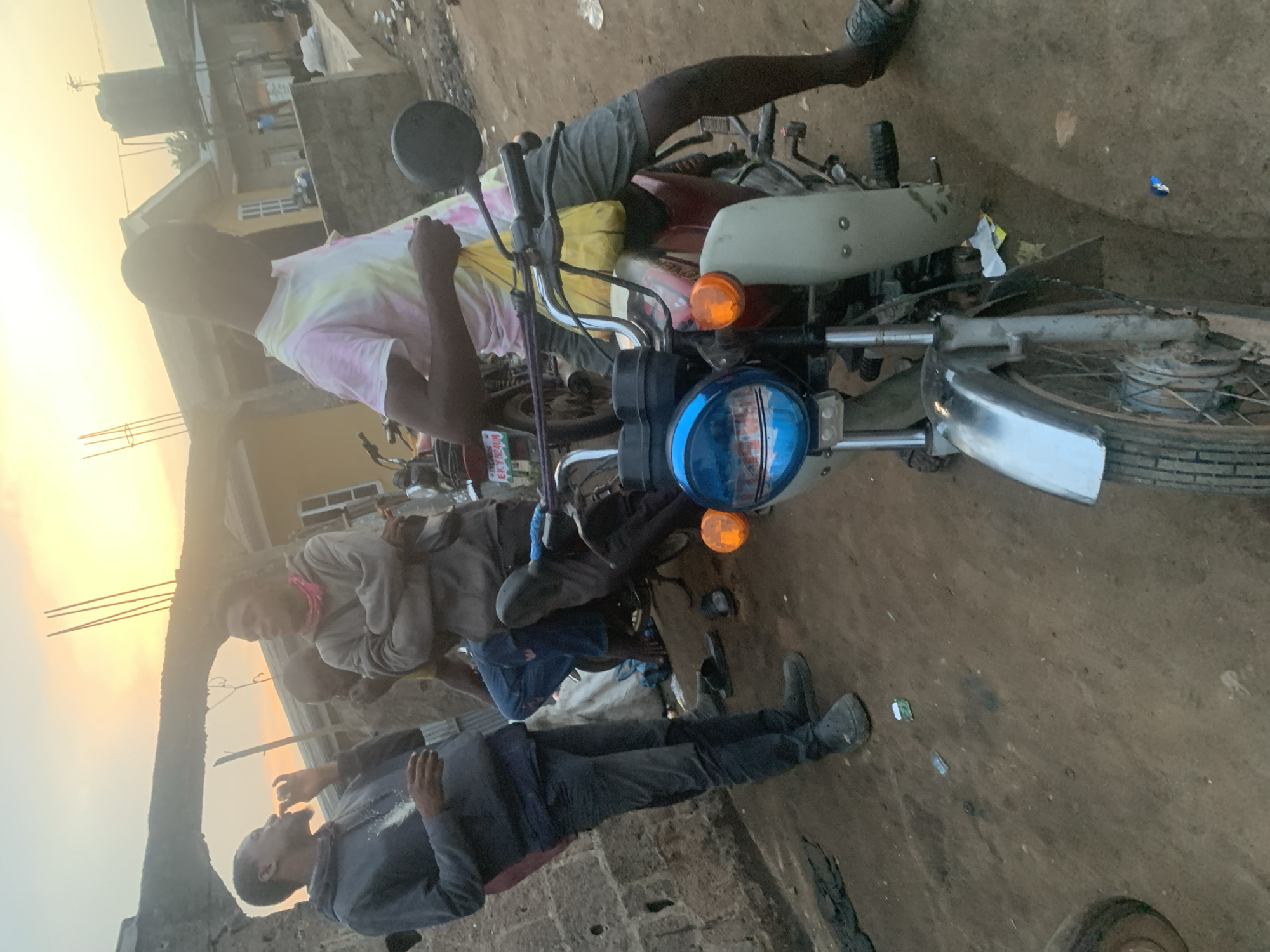 In Shagamu, Nigeria, at dusk, four young men are gathered around a red motorcycle in an outdoor, dusty area. One man on the far left wears a dark gray hoodie and dark pants. Next to him, another man in a gray hoodie with a pink bandana around his neck stands with his arms crossed. Behind them, a third man in a yellow shirt is partially visible. The man closest to the camera, wearing a tie-dye t-shirt and gray shorts, is seated on the motorcycle. He holds a yellow bag in his left hand. The motorcycle has a blue headlight, an orange turn signal, and a silver fender. In the background, a building with a yellow facade is visible, along with other structures and some debris on the ground. The sky is a warm orange and yellow, indicating the setting sun.