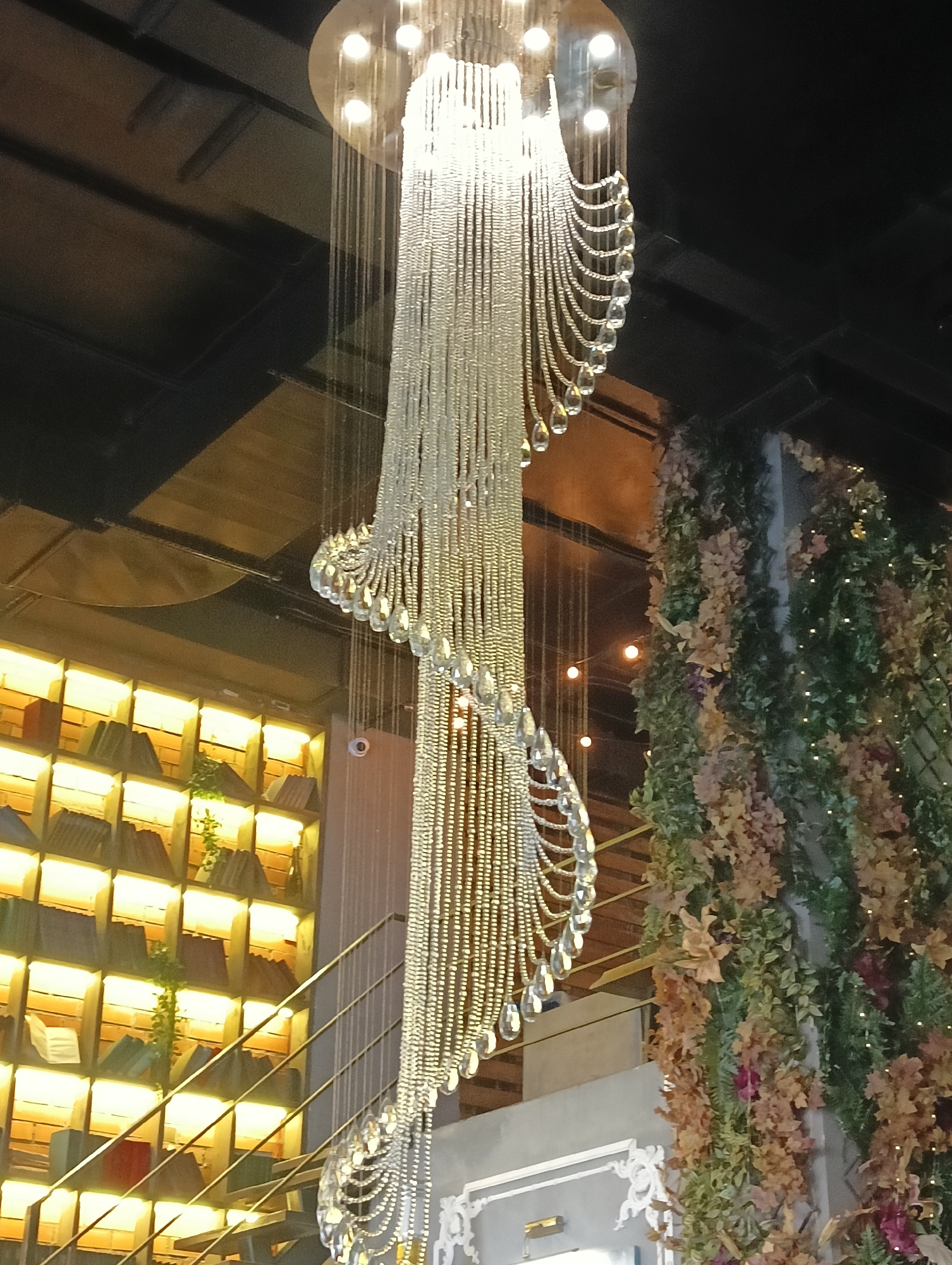 The image shows a decorative chandelier in an indoor setting. The chandelier is made of many strings of small beads and crystals, with larger crystal drops hanging at intervals. It is illuminated by several bulbs at its top. To the left of the chandelier is a wall with a shelving unit filled with books, each section of the shelving unit is lit from behind. To the right of the chandelier is a wall covered in artificial greenery and flowers, with small fairy lights interspersed. A metal railing and stairs are partially visible in the foreground. The ceiling above appears to be made of dark beams. The location is not Saint Petersburg, Russia.