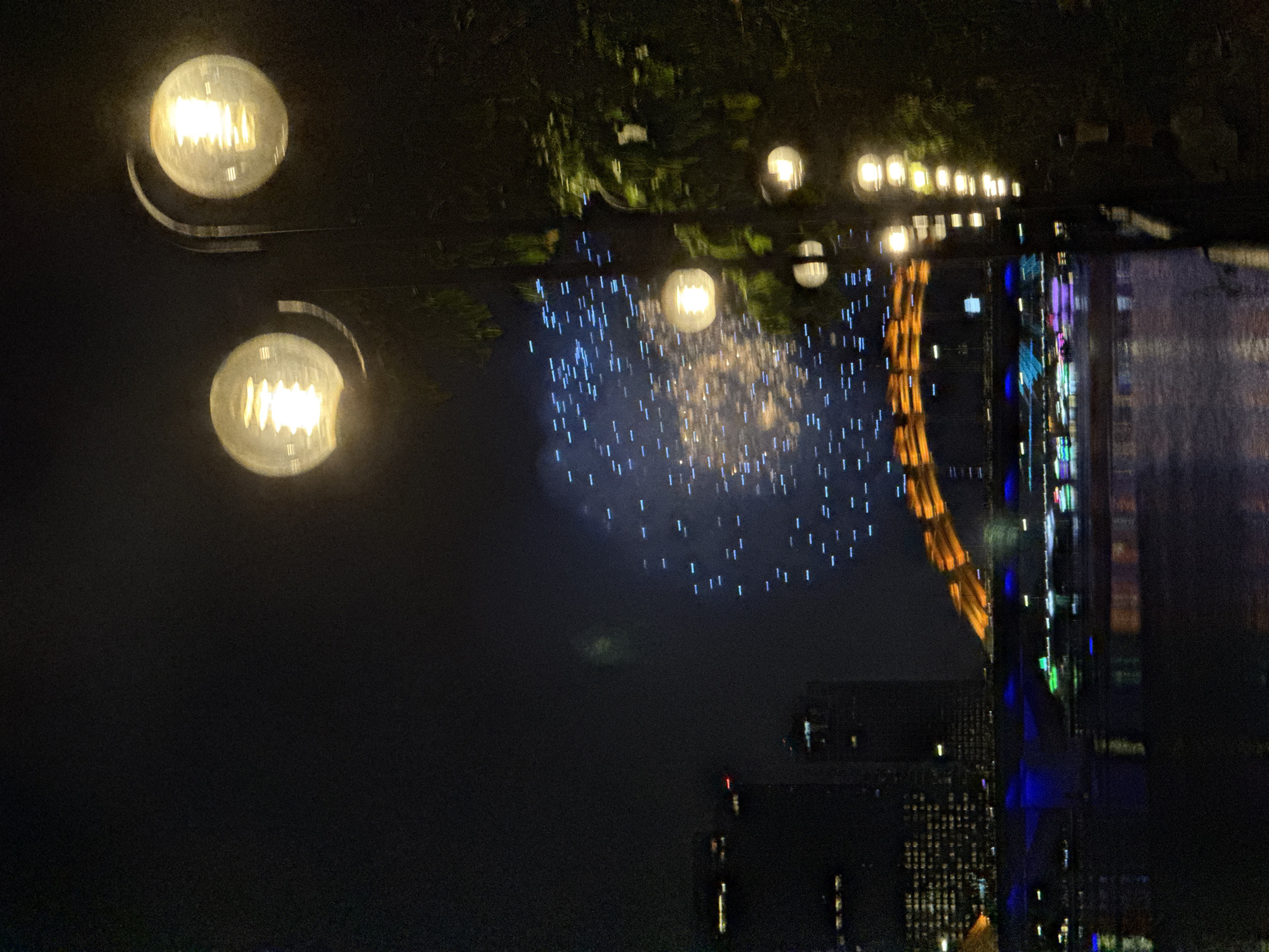 The image depicts a nighttime scene in Đà Nẵng, Vietnam. Foreground elements include two illuminated street lamps with round bulbs. The background shows a fireworks display in the sky, reflecting on a body of water, potentially the Hàn River. The skyline features several tall buildings, some lit with colorful lights. The reflection in the water mirrors the skyline and the fireworks. The environment is dark, with the primary sources of light being the street lamps, fireworks, and building illuminations.