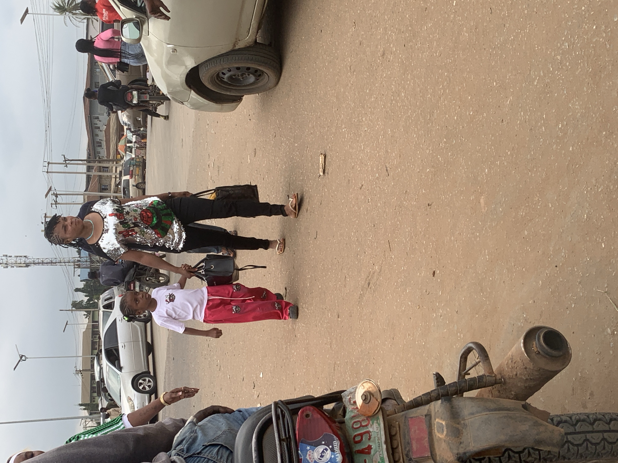 In Shagamu, Nigeria, a woman with braided hair, wearing a sequined top and black pants, walks hand-in-hand with a young girl. The girl is wearing a white t-shirt and red pants with a white drawstring. Both are holding handbags. They are walking on a dirt road in what appears to be a busy urban area, with cars, motorcycles, and buildings visible in the background. A motorcycle is parked on the left side of the frame, and another person is seen walking behind the woman and child. The scene suggests everyday life in a Nigerian city.