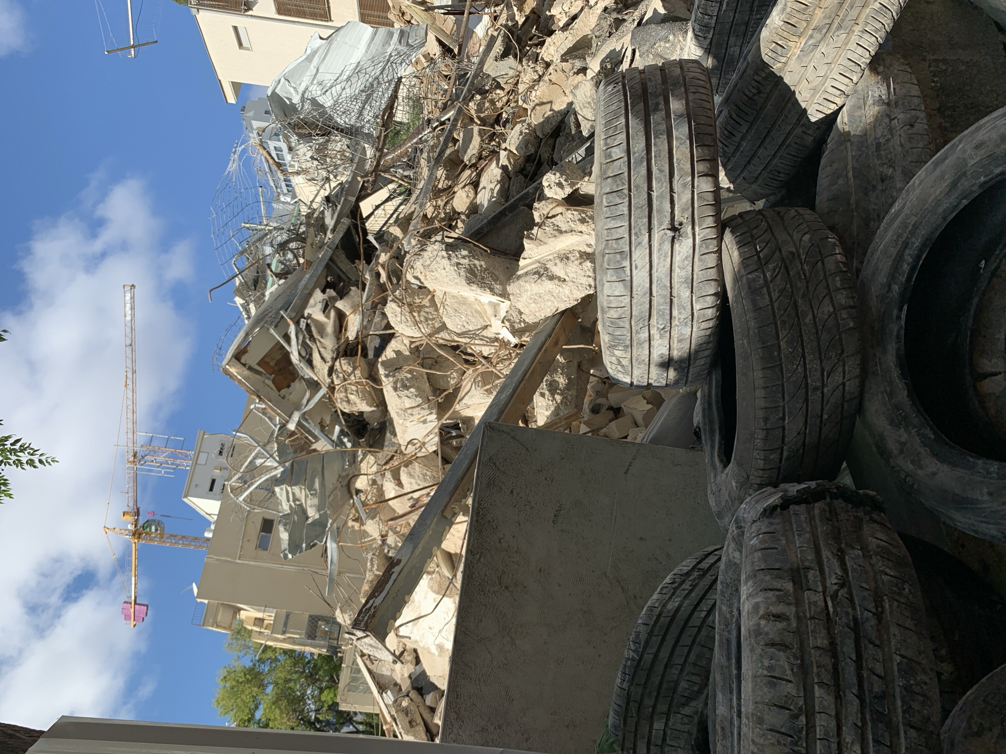 The image shows a scene of destruction, likely in Tel-Aviv, Israel, with debris from a collapsed or demolished building dominating the center and left side. A yellow construction crane is visible behind the rubble, suggesting ongoing work or demolition. A pile of used tires is stacked on the right side. The sky is blue with some clouds. The overall environment appears to be an urban construction or demolition site. No people are visible.