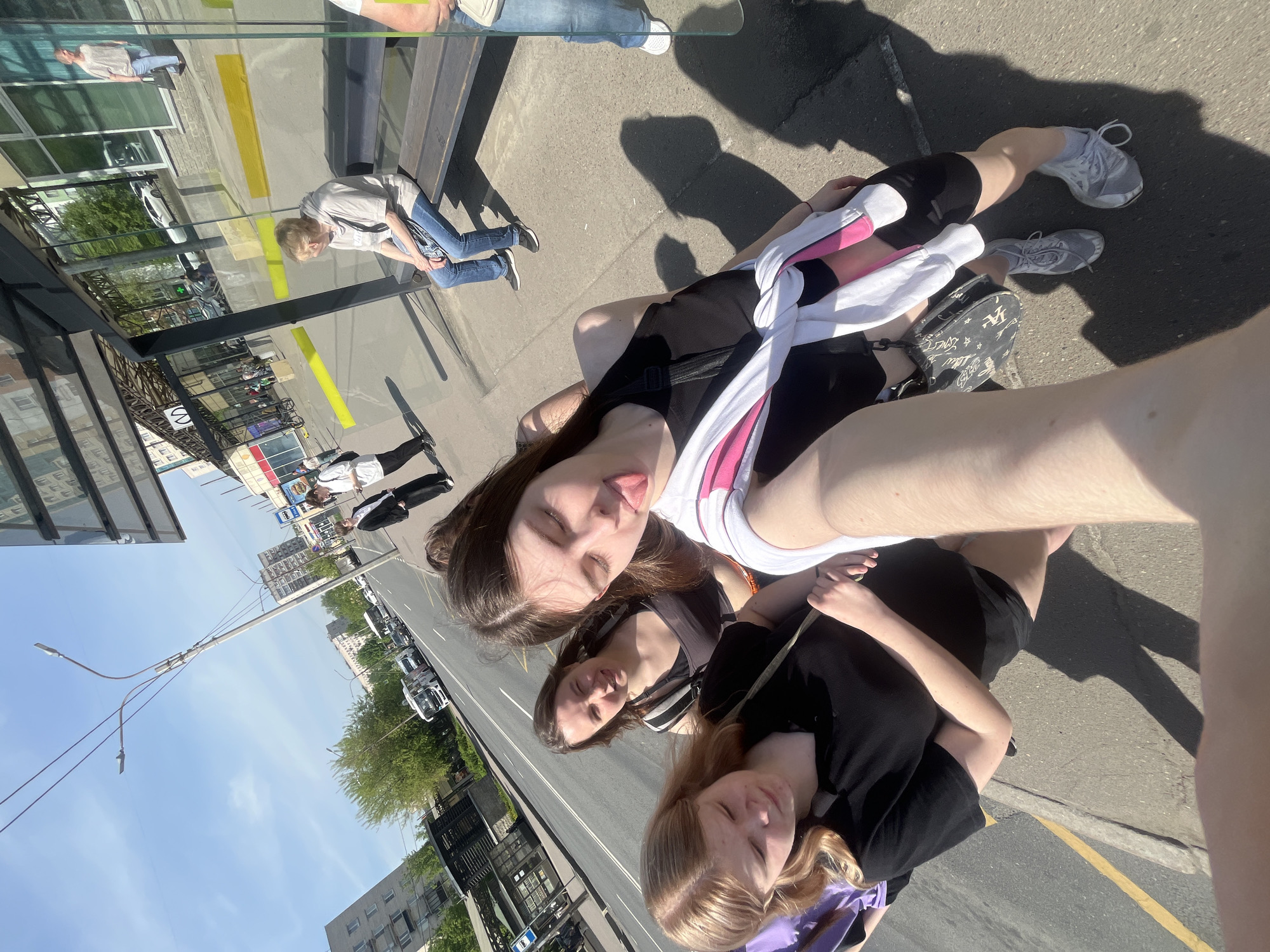 Three young women are posing for a selfie on a sunny day, standing next to a bus stop in what appears to be a city street. The background shows buildings and trees, suggesting an urban area. In the upper portion of the frame, a street light pole is visible. Reflections in the bus stop glass reveal another person sitting on a bench and passersby. The location is most likely Saint Petersburg, Russia, as suggested by the architecture, street signs, and overall style of the urban environment.