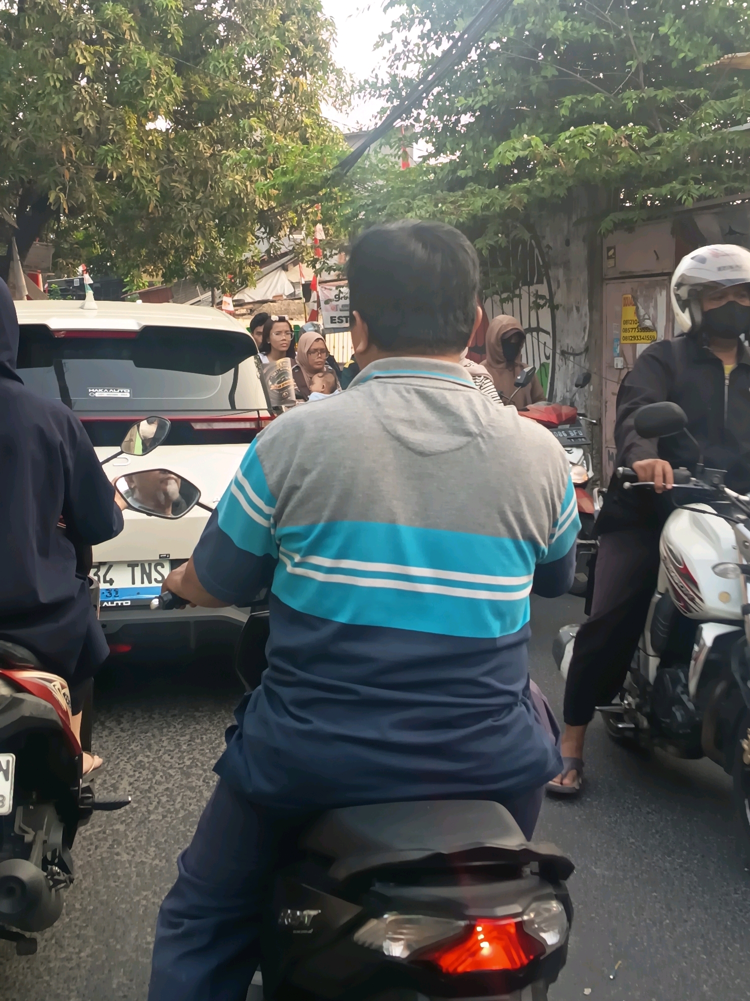 The image captures a street scene in Tangerang, Indonesia, likely during daytime traffic. In the foreground, a person is riding a motorcycle, seen from behind. They are wearing a gray and blue striped shirt and dark blue pants. The motorcycle's brake light is illuminated, indicating it is stationary or slowing down. To the left, another person on a motorcycle is partially visible, wearing a dark blue hooded jacket.
In the middle ground, a white car is visible, with its rear window slightly open. Several people are inside the car, including women wearing hijabs. One of them is holding a baby. A person on a motorcycle behind the car is wearing a hooded jacket and appears to be looking at their phone.
Further to the right, another motorcycle is stopped, with its rider wearing a white helmet and a black mask. They are wearing a black jacket and dark pants. The rider's hand is on the handlebar.
The environment appears to be an urban setting with trees lining the street and buildings in the background. There is evidence of traffic, with multiple vehicles present. The road surface is asphalt. Some posters or signs are visible on a wall to the right. The overall impression is a congested street during normal daily activity in Tangerang.