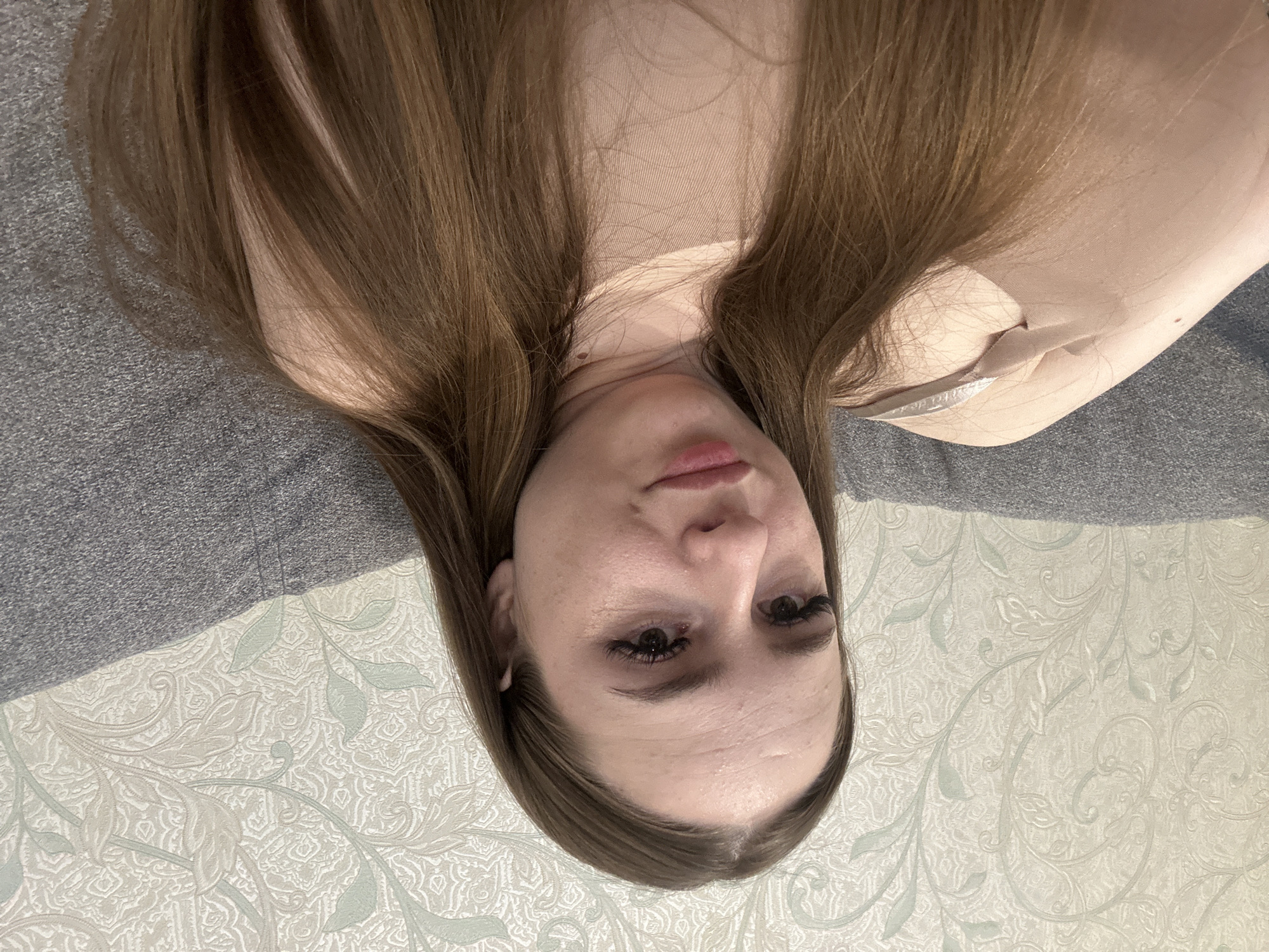 A person with brown hair is lying on a gray surface. The background is a patterned wall. The location is Unknown, Unknown.