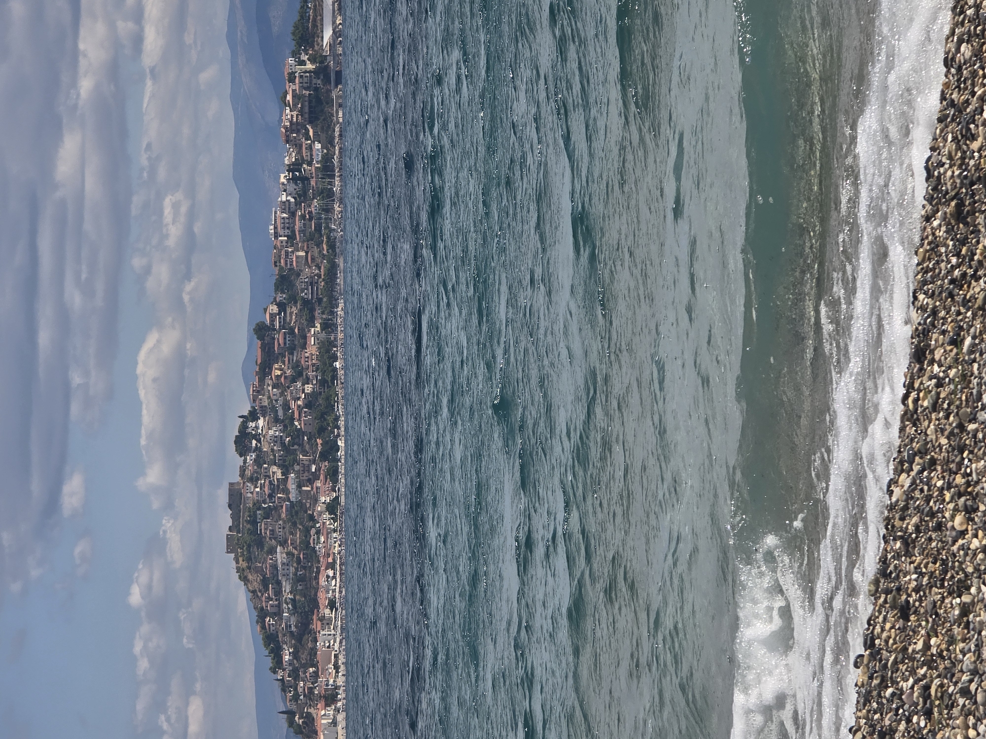 The image shows a view of the town of Astros, Greece, from the sea. In the foreground, there is a pebble beach with gentle waves lapping the shore. The water is a deep blue-green color. In the middle ground, the town of Astros is visible, built on a hillside overlooking the sea. The houses are white with red tile roofs, and there is a castle on the highest point of the hill. In the background, there are mountains. The sky is blue with scattered white clouds.