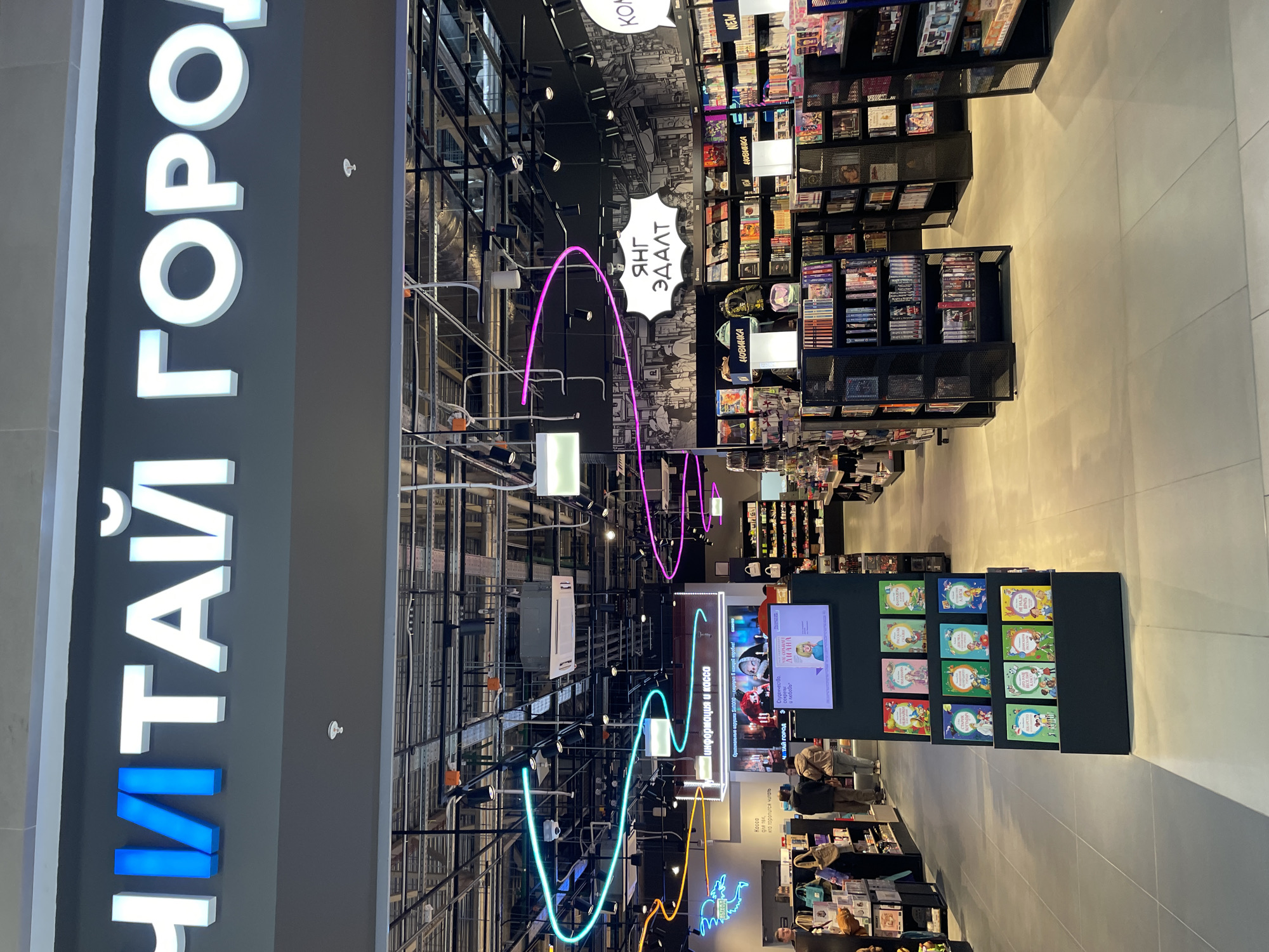 The image depicts the interior of a "Chitai-Gorod" bookstore, as indicated by the large signage in Russian, meaning "Read City." The shot is taken from an angle, looking down the aisle. The store features black metal shelving stocked with books, movies, and other merchandise. There are neon lights in purple, blue, and yellow, hanging from the ceiling along the aisle. A sign in the shape of a speech bubble with text in Russian is hanging down. There are several people, including shoppers, browsing the lower shelves and a cashier visible near the bottom of the image. The scene has a modern and well-lit atmosphere. The location is in the city of Kazan, Russia.