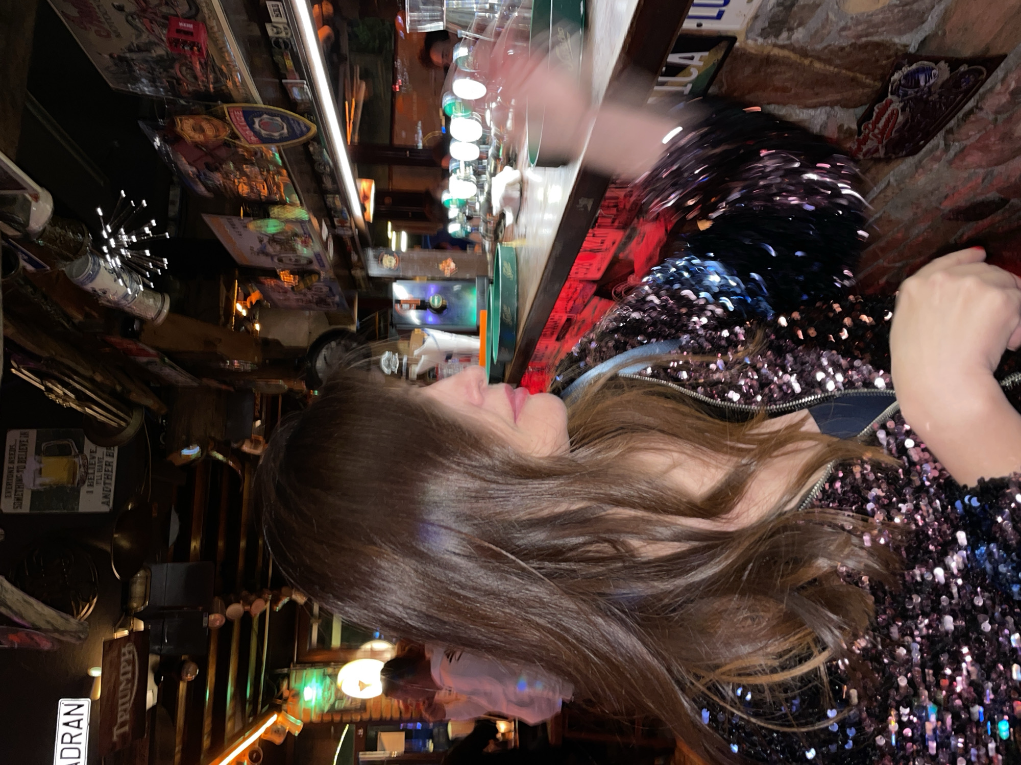 The image shows a woman with long brown hair, wearing a sequined jacket, seated at a bar in a dimly lit establishment. The bar is adorned with various decorations including mugs, lights, and framed posters, one of which says "TRIUMPH". The scene is suggestive of a bar in Budva, Montenegro.