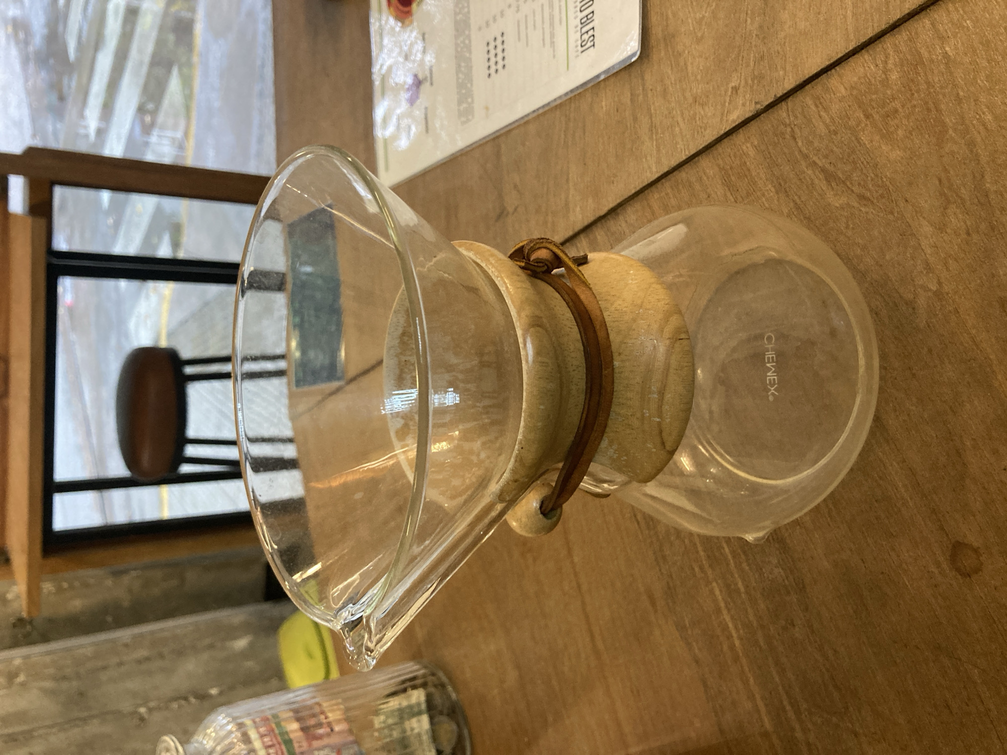 The image displays a clear glass Chemex coffee maker resting horizontally on a wooden table. The coffee maker features a light-colored wooden collar secured by a brown leather tie with a small bead. It appears to be empty and clean.
No people are visible in the frame. Other objects include a partially visible menu or document in the upper background, which is also resting on the wooden table. Near the bottom left, part of another glass bottle or jar with a colorful label is discernible. The setting appears to be an indoor environment, likely a coffee shop or café, as suggested by the Chemex and menu.
In the background, a large window is visible, through which faint reflections of an outdoor scene can be seen, possibly indicating an overcast or rainy day due to the diffused light and streaks on the glass. A dark-framed window section and a dark brown stool are also visible through this window. The table surface is made of light to medium brown wood with a visible grain. A subtle water ring or stain is present on the table to the right of the Chemex's base.
No specific activity is taking place; the Chemex seems to be either on display or waiting to be used. The visible text "CHEMEX" is clearly printed on the side of the coffee maker. On the menu in the background, the partial text "O BEST" can be read. The image itself is rotated 90 degrees counter-clockwise.