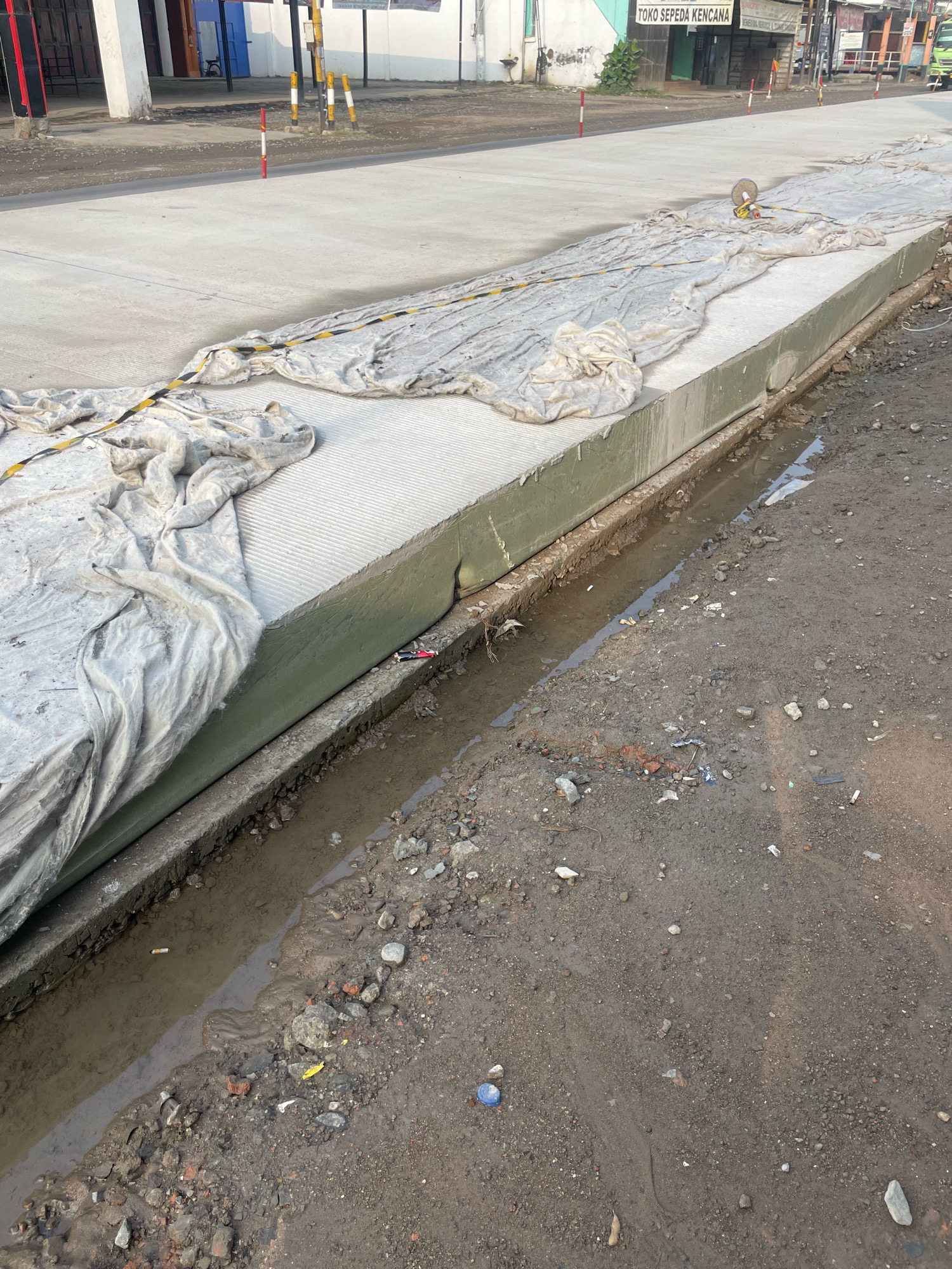 A long, concrete construction project is underway, with a section of it covered by a white tarp. The tarp is draped over a raised concrete structure. There's a narrow ditch filled with murky water running alongside the concrete. The ground is a mix of dirt, gravel, and scattered debris, including cigarette butts and small pieces of brick. In the background, buildings and a street are visible, with a few traffic poles and signs. The sky is not visible in the frame.