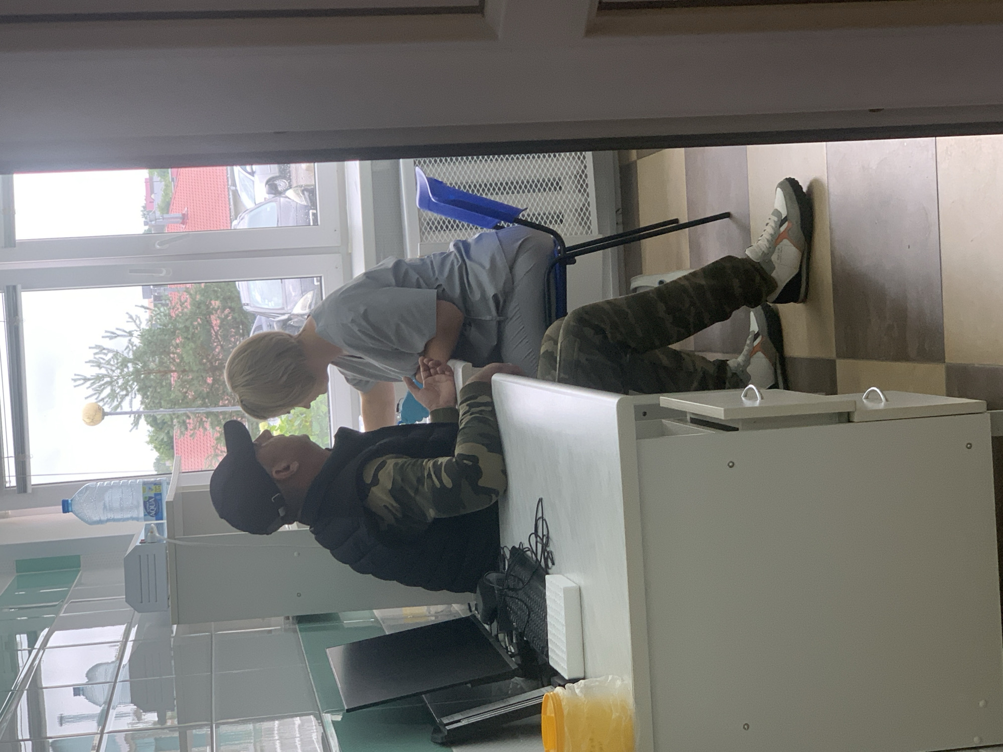 The image depicts an indoor scene, likely a medical or laboratory setting, with two individuals. A man wearing a black baseball cap, a dark vest over a camouflage-patterned long-sleeved shirt, and camouflage pants is seated at a white desk. His left arm is resting on the desk, and his right hand is being held by a woman. The woman, with short blonde hair, is wearing light blue medical scrubs and is seated on a blue chair facing the man. She appears to be looking at his hand.
On the desk in front of the man are a computer monitor, a keyboard, a mouse, a white rectangular object that could be a sample rack or a small device, and a clear plastic container with a yellow lid. Behind the desk, a large window reveals an outdoor scene with trees and buildings. The room has tiled walls and a tiled floor. A white cabinet is visible to the left of the desk, and a tall blue chair is positioned to the right of the desk. The presence of medical attire and the desk setup suggests a healthcare or research environment. The location within the city of Kapyĺ, Belarus, is not explicitly indicated by any visual cues in the image.