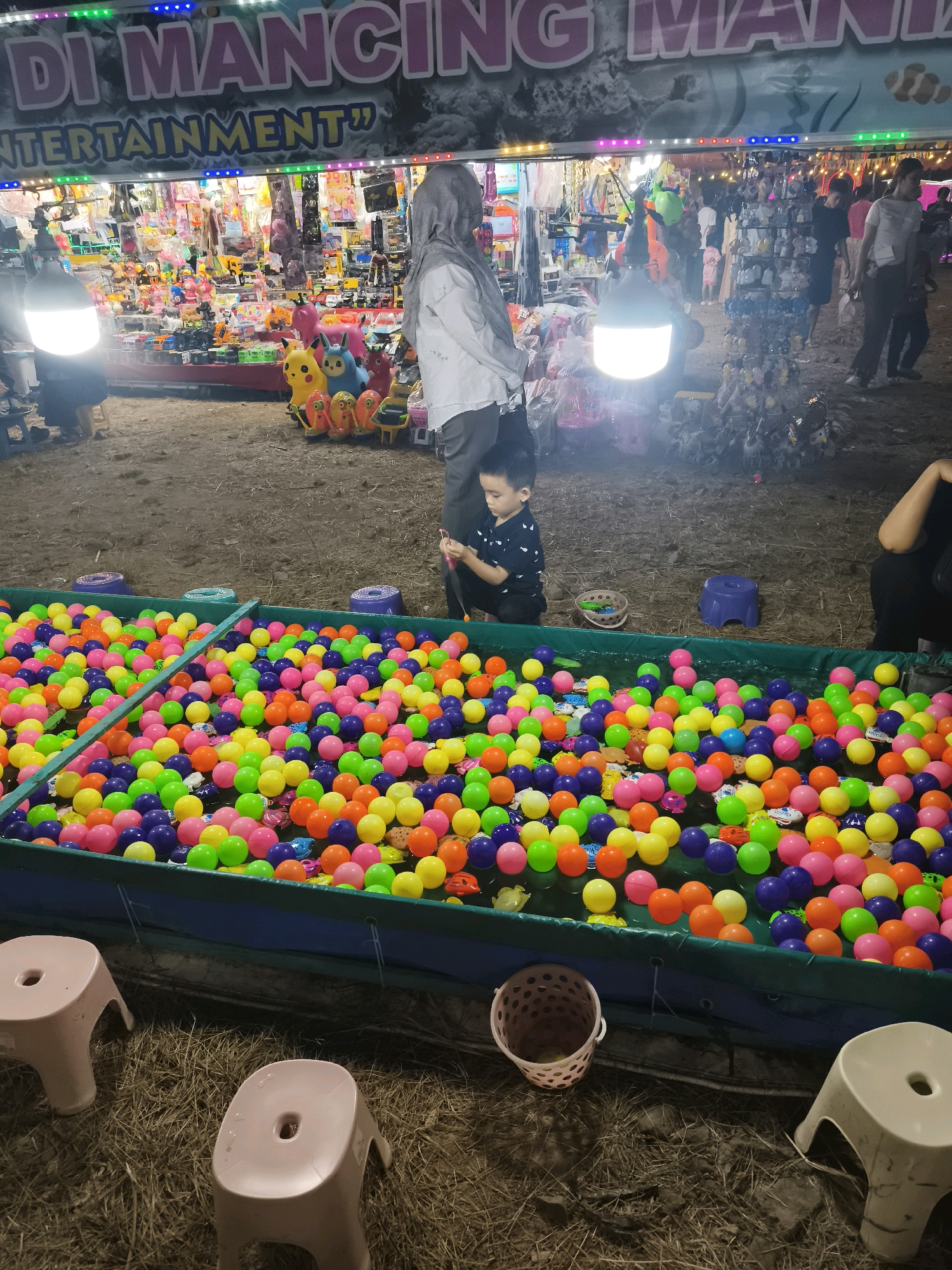 The image depicts an outdoor carnival or fairground scene, likely in Palembang, Indonesia. In the foreground, a large, shallow pool is filled with numerous brightly colored plastic balls, predominantly pink, yellow, green, and purple. Several small toy fish and other miniature toys are floating amongst the balls. Two small, light-colored plastic stools are visible to the left and bottom right of the ball pool, placed on a ground covered with dry straw or hay. A small, pink plastic basket with holes is positioned near the bottom center of the frame.
In the middle ground, a young boy wearing a dark patterned shirt is kneeling beside the ball pool, actively engaged with a fishing rod-like toy, seemingly attempting to catch one of the floating toys. To the left of the boy, a woman wearing a grey hijab and a white shirt is standing, also looking towards the ball pool area.
Further in the background, a bustling stall or booth is brightly lit with colorful lights. The stall is stocked with an assortment of toys and carnival merchandise, including stuffed animals, toy cars, and other novelty items. Several people are visible browsing or walking past the stall, indicating a lively atmosphere. A large banner with pink lettering, partially obscured, reads "DI MANCING MAN" followed by "ENTERTAINMENT". This banner suggests the event is a fishing-themed game or entertainment venue. The overall environment appears to be a temporary setup, common at local fairs or festivals.