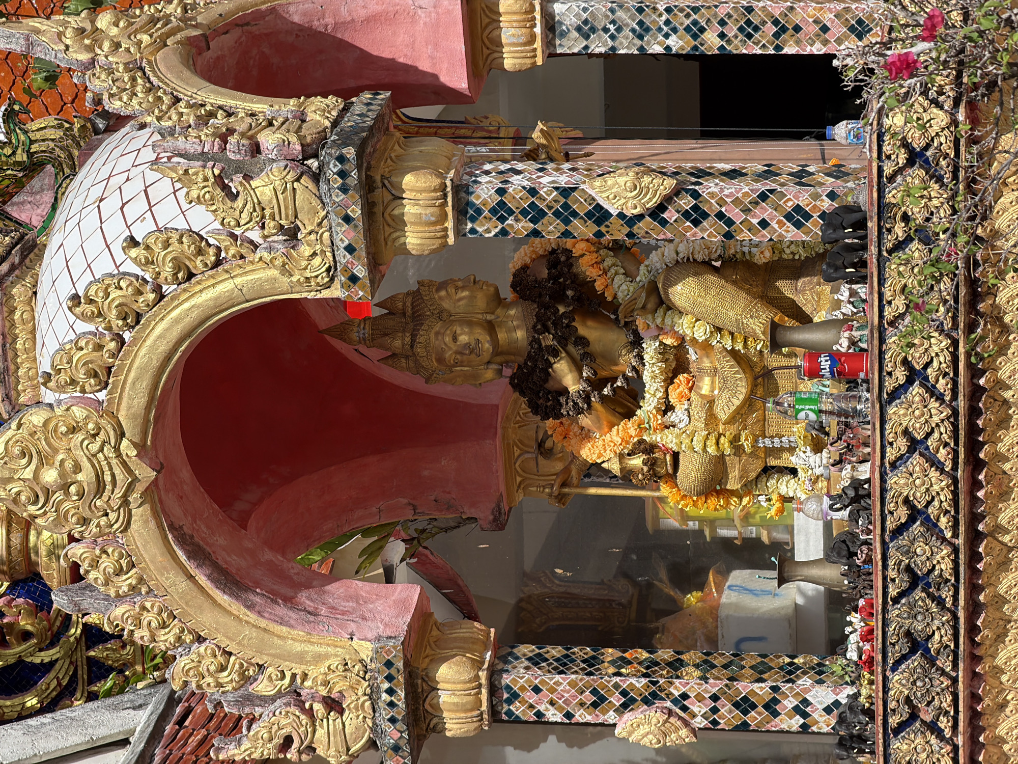 The image depicts a shrine at Baan Plai Laem Temple in Koh Samui, Thailand, featuring a large, golden statue with multiple faces, adorned with garlands of orange and white flowers. The statue is housed within an ornate, arched structure with gold-colored decorative elements and pink-painted arches. The columns supporting the structure are decorated with colorful mosaic tiles. In front of the statue, on a decorative ledge, are numerous small figurines, predominantly black elephants, along with some red and white elephant figures, and other small statues. A red can of a beverage and a green bottle of water are also visible on the ledge. The background shows a glimpse of the temple's architecture, including a white dome-like structure. Lush green foliage with pink flowers is in the foreground, framing the lower part of the shrine. The scene is bathed in bright sunlight, casting distinct shadows.