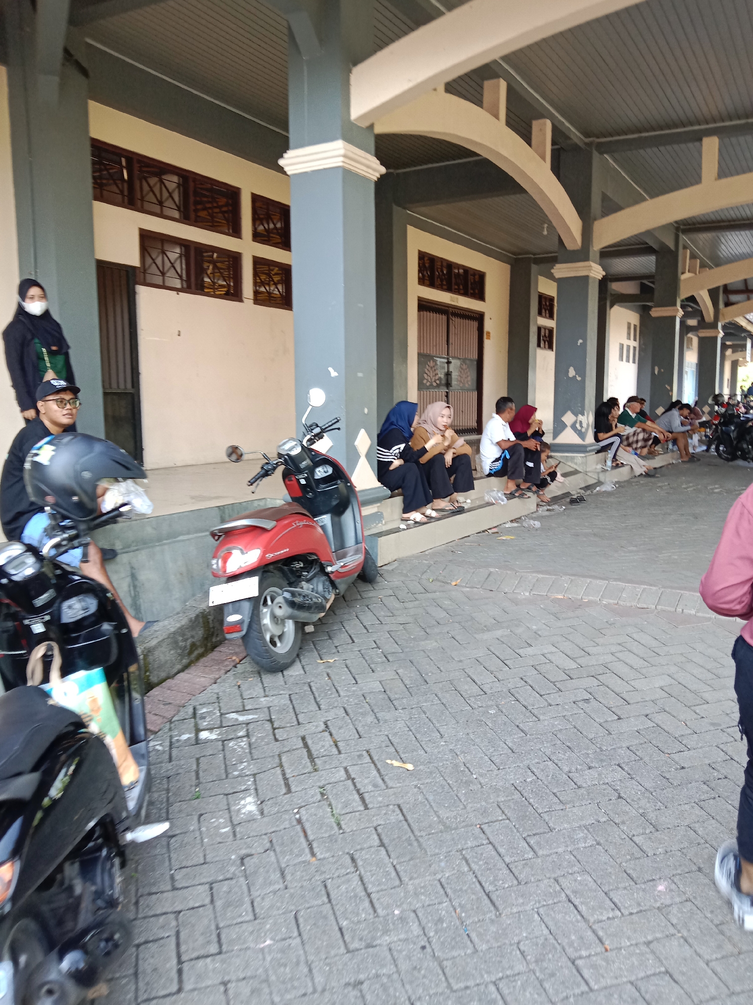 This image captures a scene outside what appears to be a building, possibly a public facility or market, in Kendal, Indonesia. Several people are seated on steps leading up to the building, while others are standing. Two scooters are parked on the paved walkway in the foreground. The building has a covered walkway supported by columns, and windows with decorative grilles. The overall environment suggests a warm, possibly sunny day.