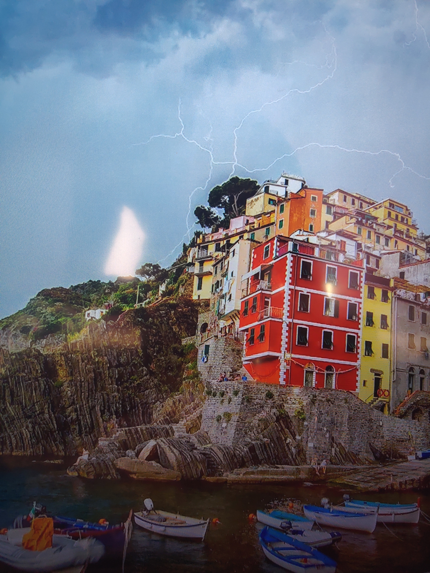 The image depicts the coastal village of Riomaggiore in Cinque Terre, Italy. The village is built on a rocky hillside that slopes down to the sea. The buildings are colorful and stacked on top of each other. Several boats are in the harbor. Lightning is visible in the sky.