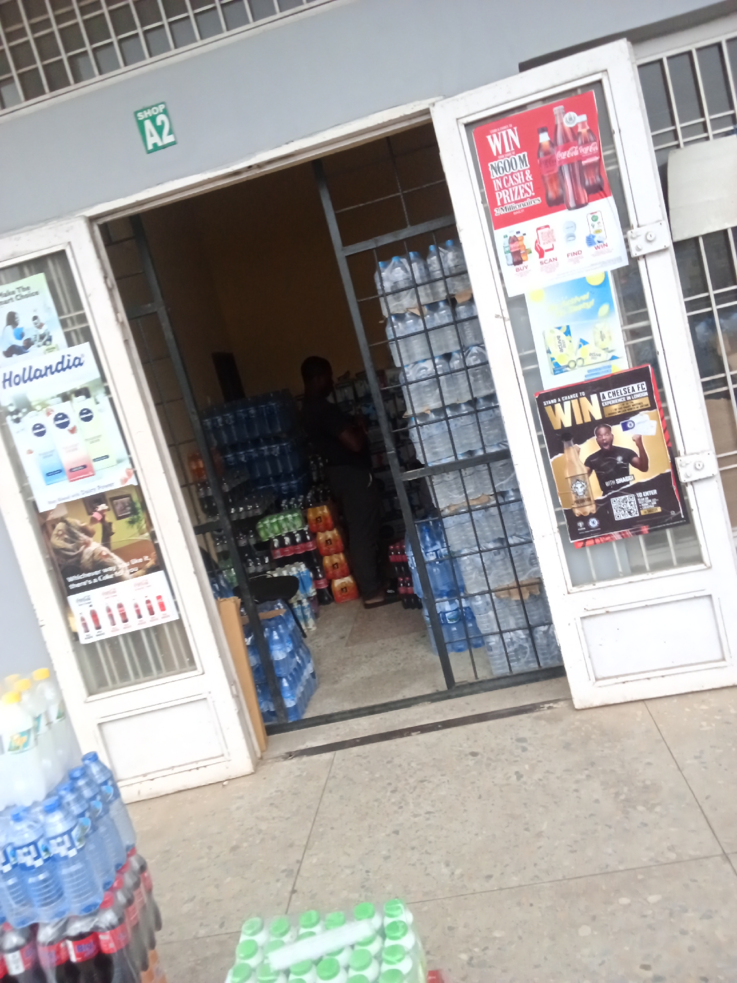 The image depicts the entrance of a small shop, designated as "SHOP A2", located in Jos, Nigeria. The shop appears to be a convenience store or a small supermarket, with a large display of bottled water and other beverages visible both inside and outside the entrance. A man in a dark t-shirt and trousers is standing inside the shop, seemingly attending to customers or stocking shelves. Several promotional posters are displayed on the glass doors of the shop, advertising various products and promotions, including a "WIN N600M IN CASH & PRIZES!" campaign by Coca-Cola and a "STAND A CHANCE TO WIN A CHELSEA FC EXPERIENCE IN LONDON" promotion. The floor is paved with concrete, and the overall environment suggests a commercial area in Jos.