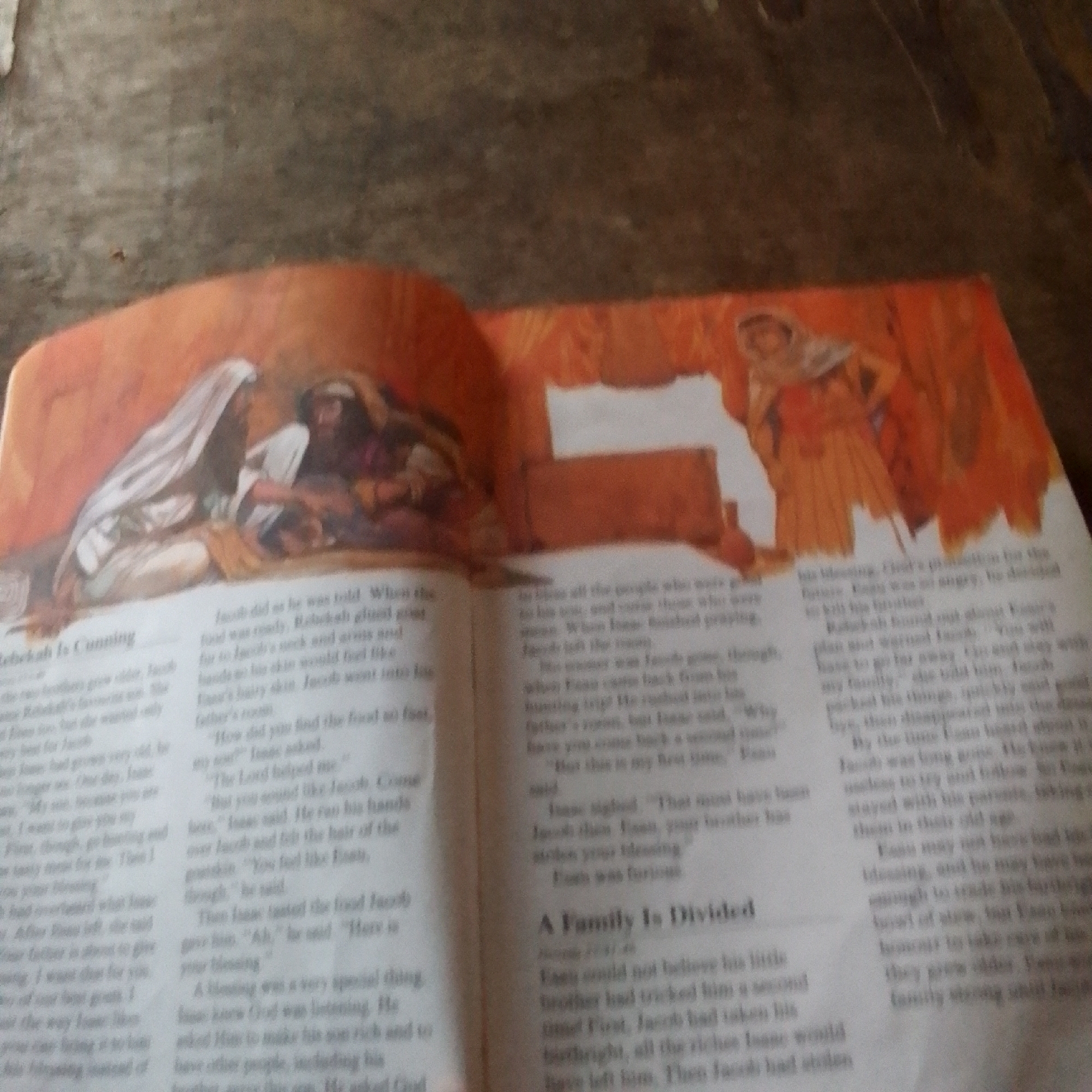 The image shows an open book with illustrations and text. The book appears to be religious or historical in nature, depicting figures in traditional attire. One illustration shows two figures, one with a white head covering, interacting with another seated figure. The text is in English and discusses biblical characters like Rebekah, Jacob, and Esau. The overall scene is a close-up of an open book, likely on a flat surface, with an orange-toned background dominating the right side of the image, possibly suggesting the cover or another page of the book. The lighting is uneven, casting shadows and making some parts of the image less clear. There are no discernible elements that would allow for the identification of a specific city like Jalingo, Nigeria, or any forensic details related to a crime scene.